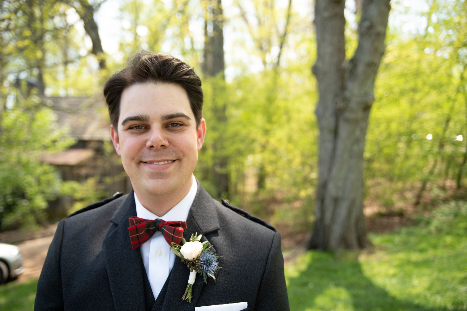 A smiling groom in a dark suit, red bow tie, and floral boutonniere poses outdoors on his wedding day.