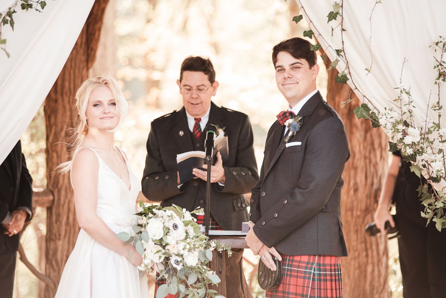 Smiling bride and groom in Scottish tartan kilt standing with officiant under floral arch at outdoor wedding.
