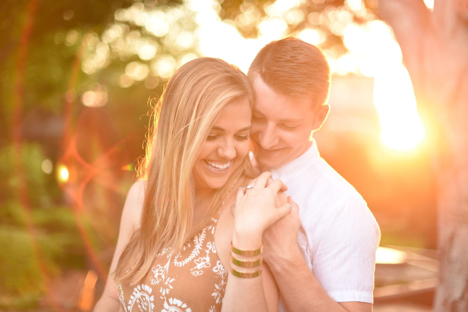 Happy couple embracing at golden sunset, bride smiling warmly in floral dress during engagement photo.