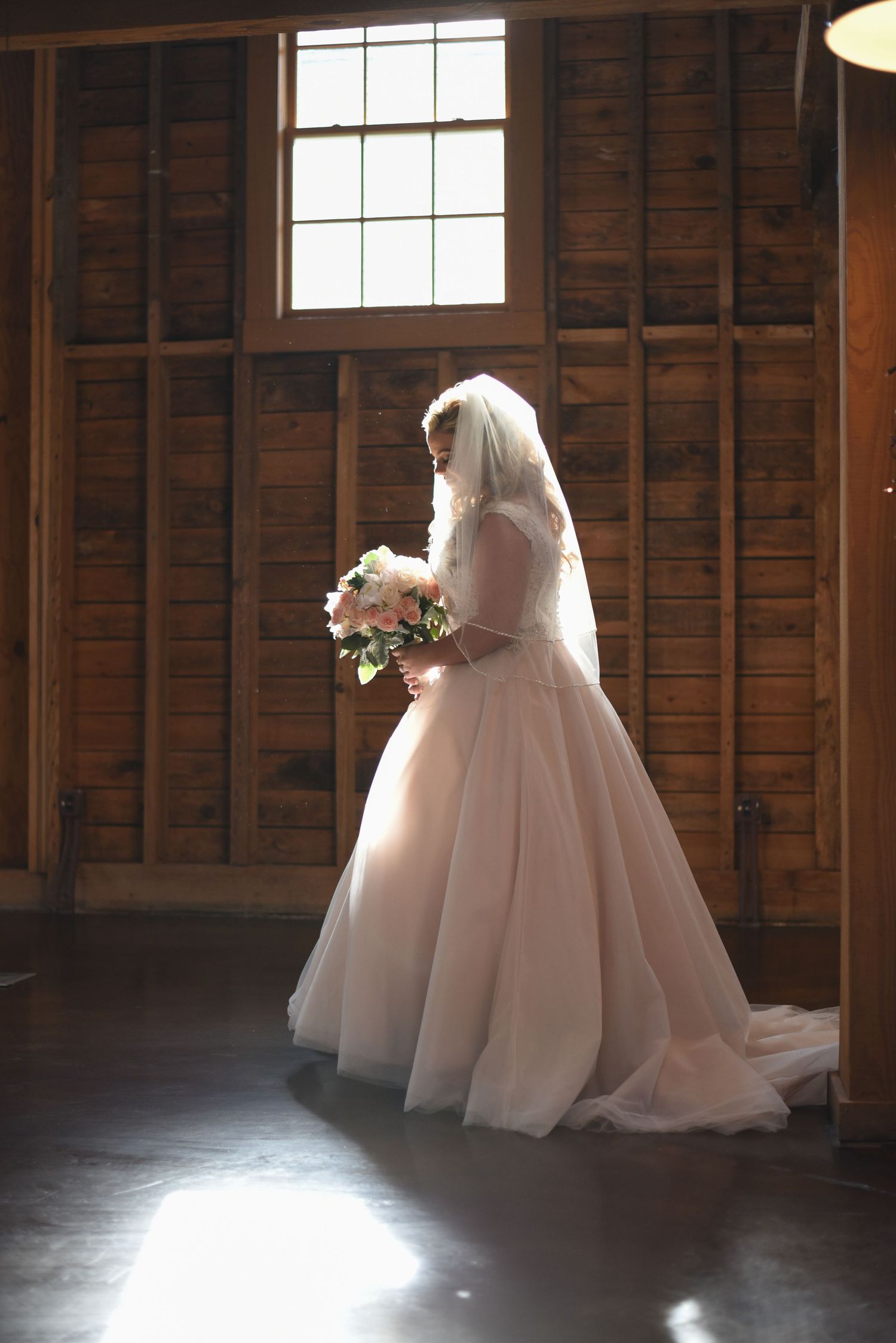 Bride in a blush ball gown and veil holds a floral bouquet in a rustic wooden barn with natural window light.