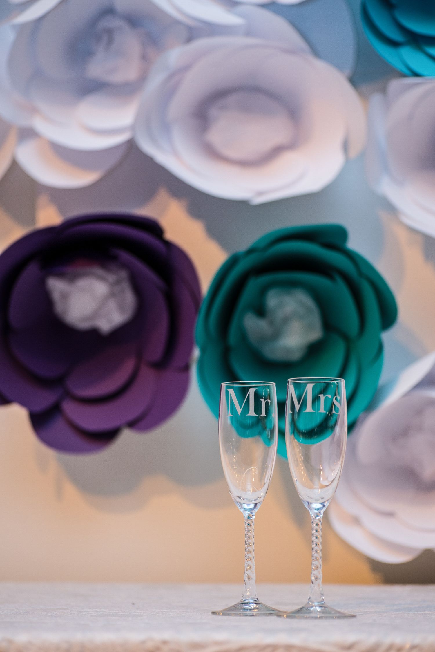 Mr. and Mrs. engraved champagne flutes displayed in front of colorful paper flower backdrop in purple, teal, and white.