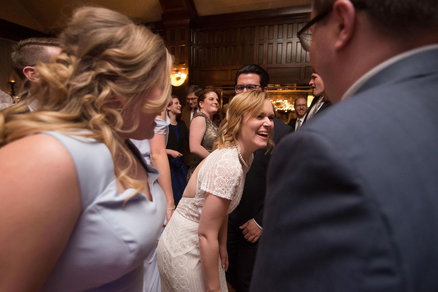 Bride in white lace dress laughing joyfully with guests on wedding reception dance floor.
