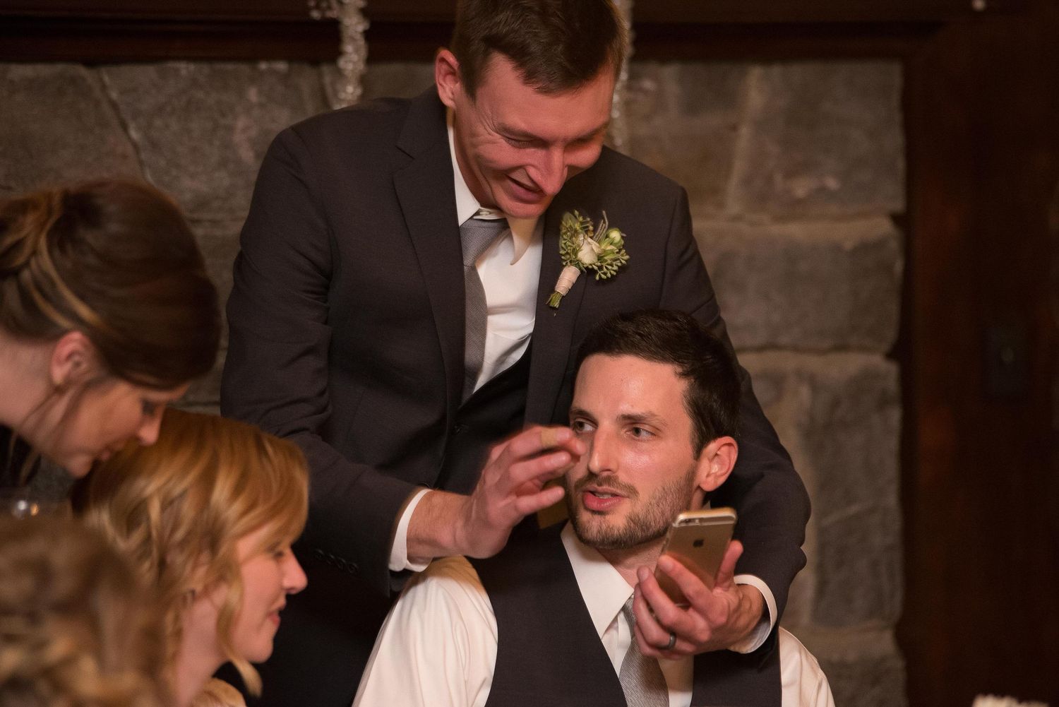 Groom in suit with boutonniere laughing with seated guest holding phone at elegant wedding reception.