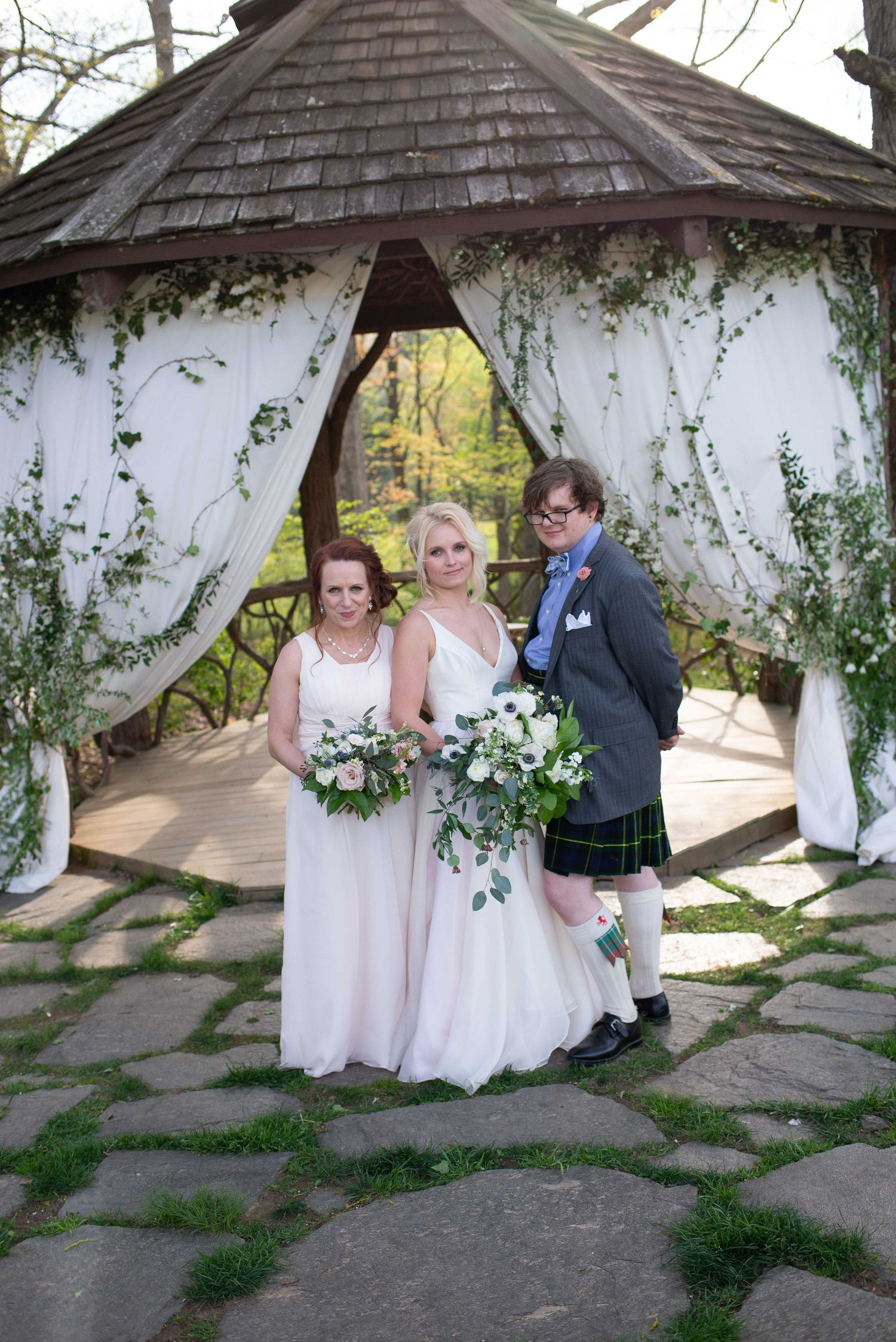 Two brides and officiant pose under a floral gazebo with white drapes at an outdoor wedding ceremony.