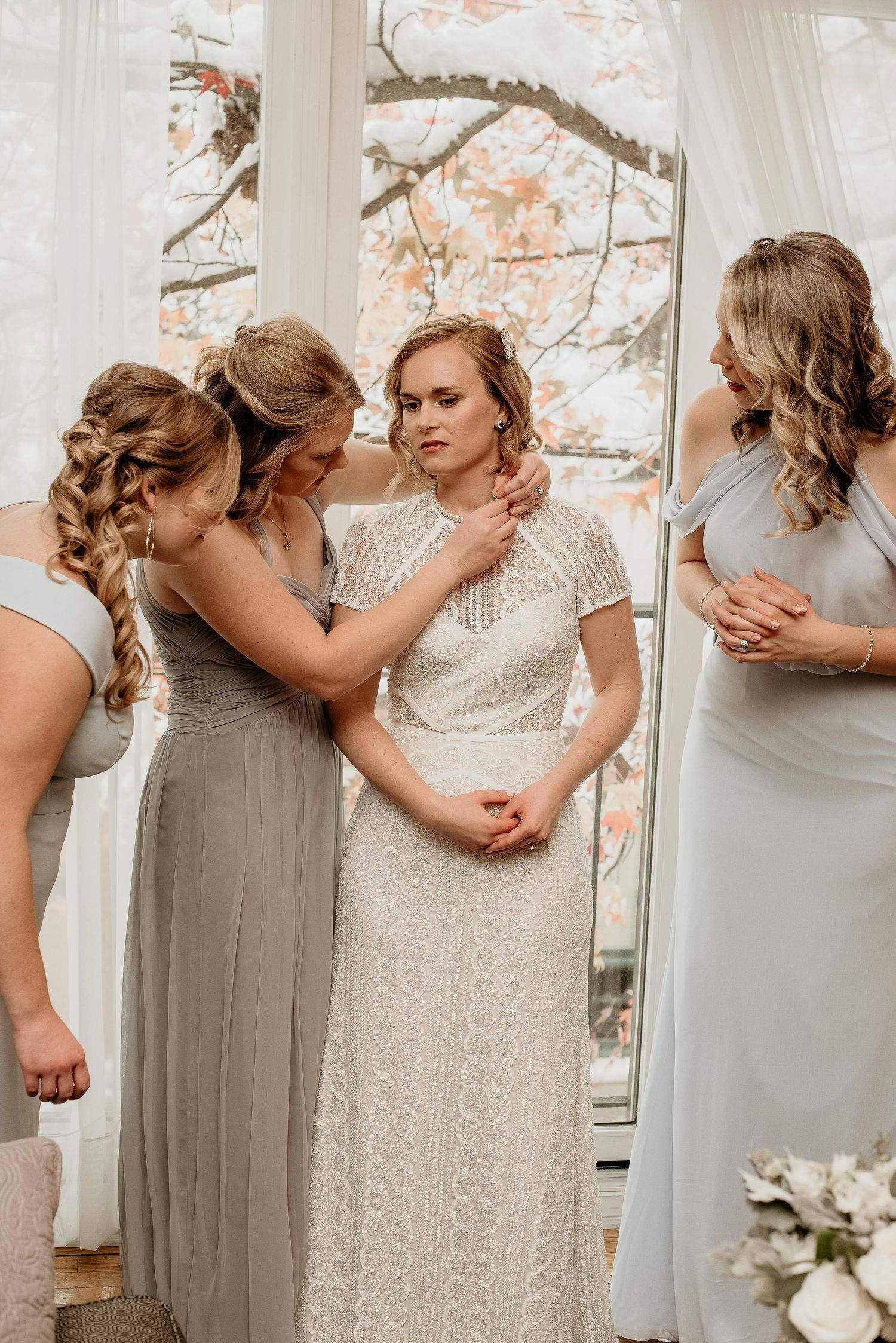 Bride in white lace gown surrounded by bridesmaids in gray dresses during wedding preparation indoors.