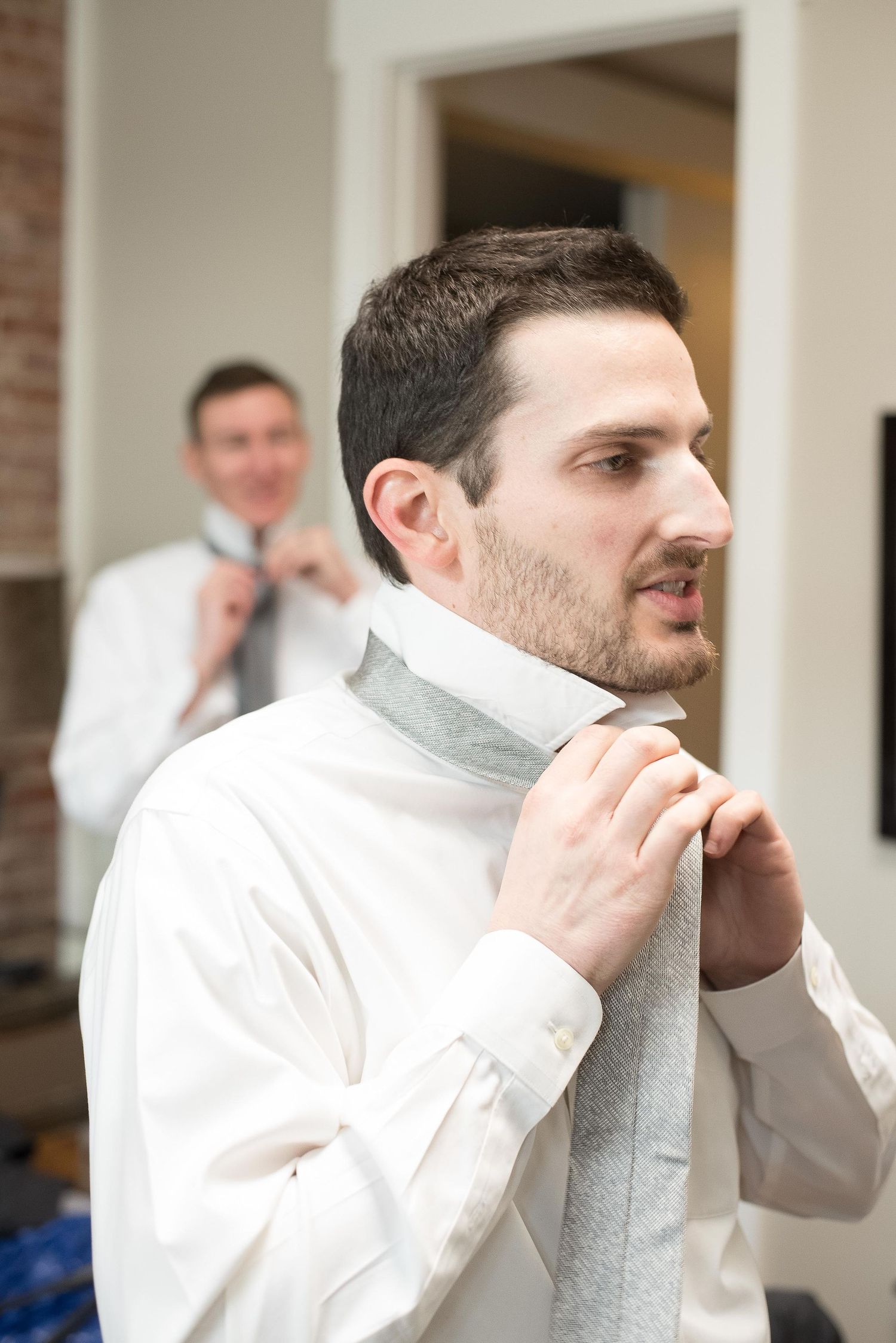 Groom adjusting gray tie on white dress shirt while another man fixes his tie in background.