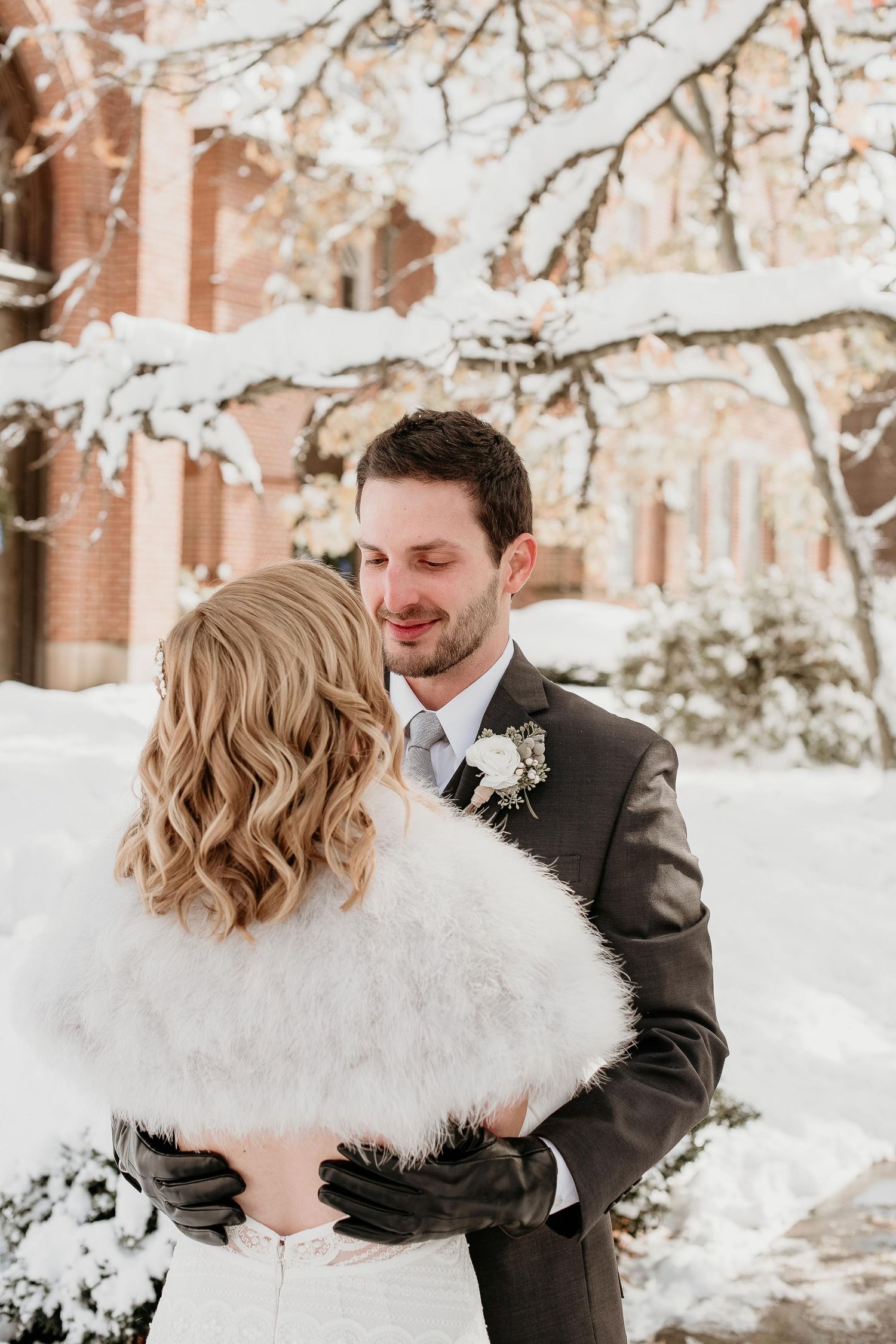 Bride and groom embrace in snowy winter setting, she wears white fur wrap, he wears dark suit with boutonniere.