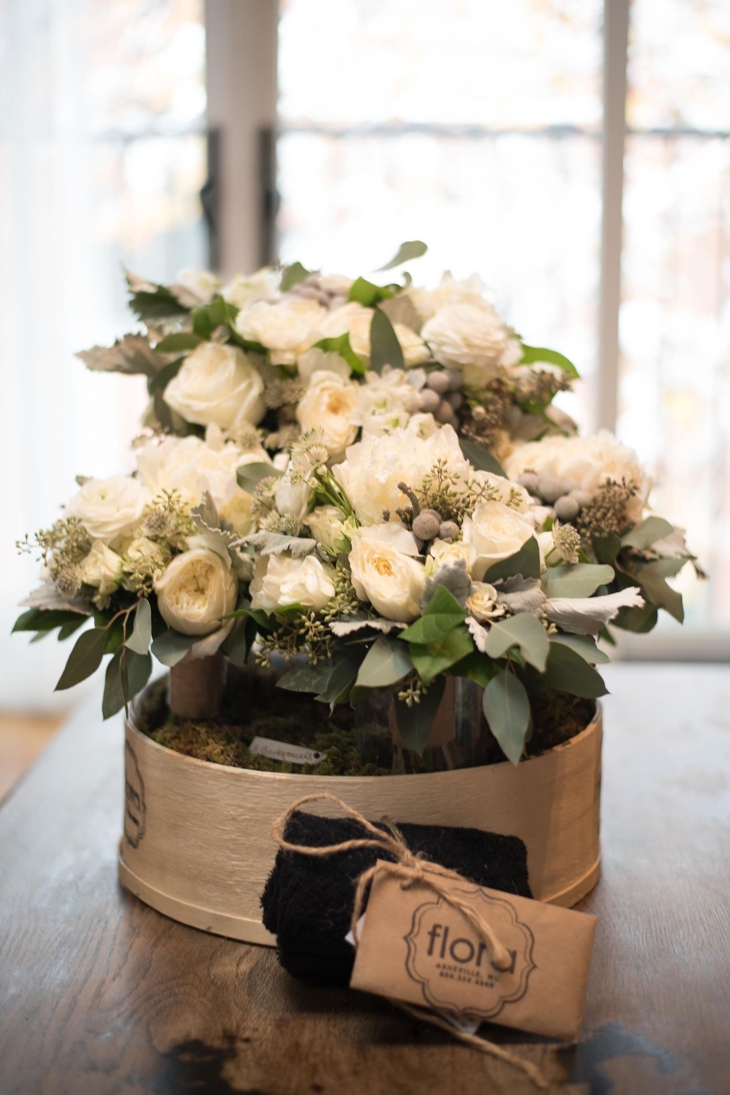 Elegant white rose and eucalyptus floral arrangement in a rustic wooden box with a Flor boutique tag on a wooden table.