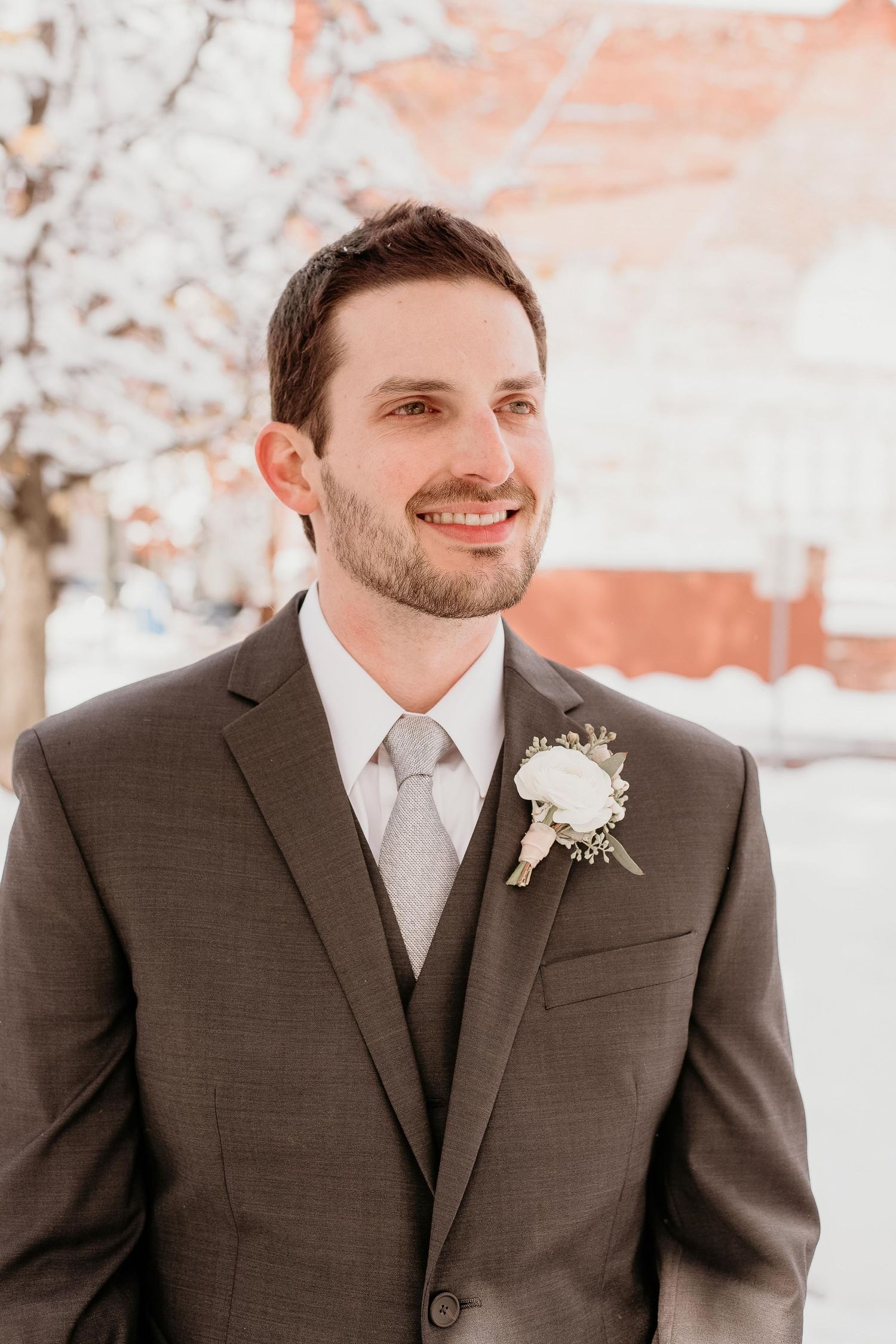 Smiling groom in gray suit with white boutonniere poses outdoors in snowy winter setting.