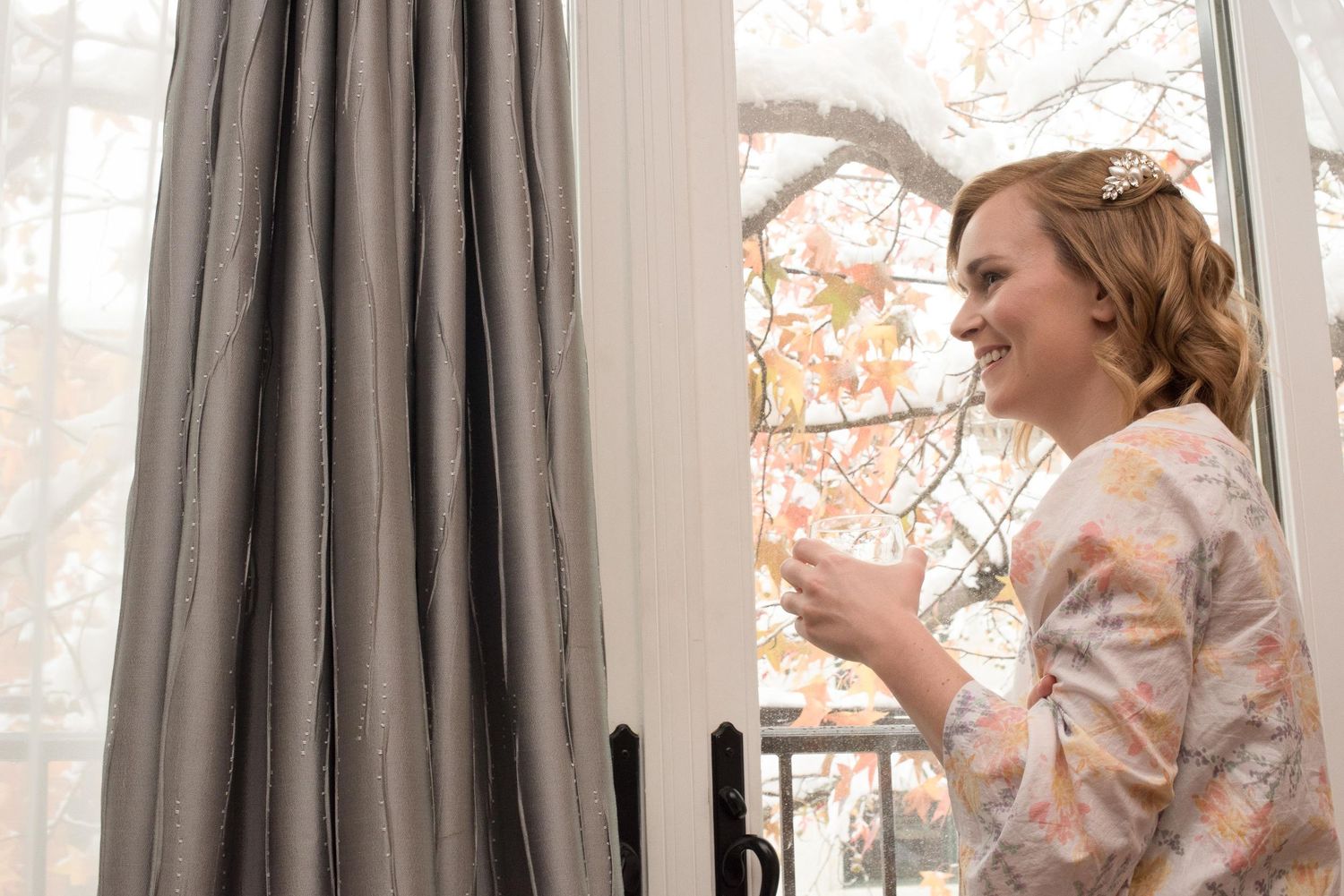 Smiling bride in floral dress holding a cup, standing by a window with gray curtains and autumn trees outside.