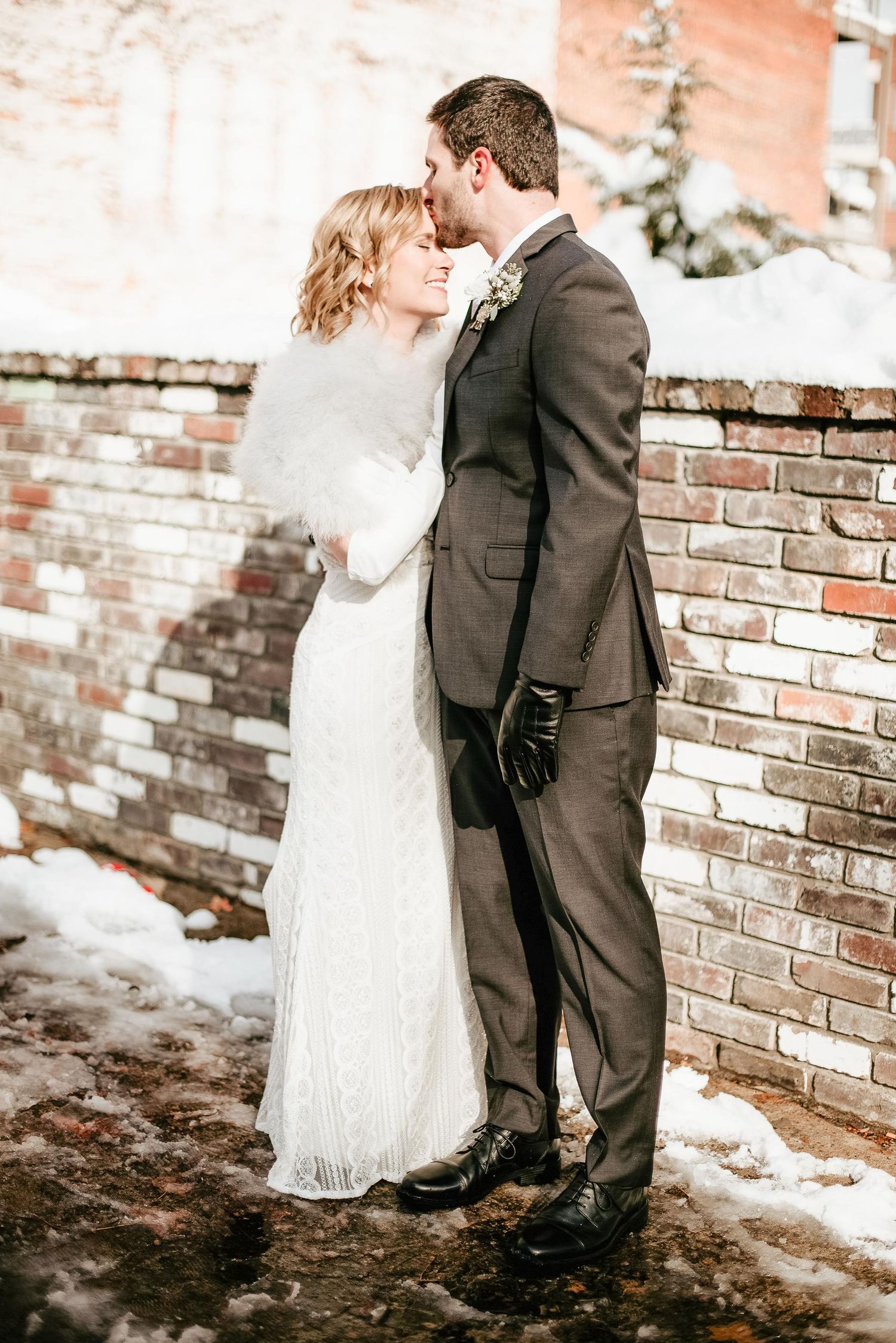 Bride and groom share a romantic winter kiss in the snow, wearing elegant wedding attire beside a brick wall.