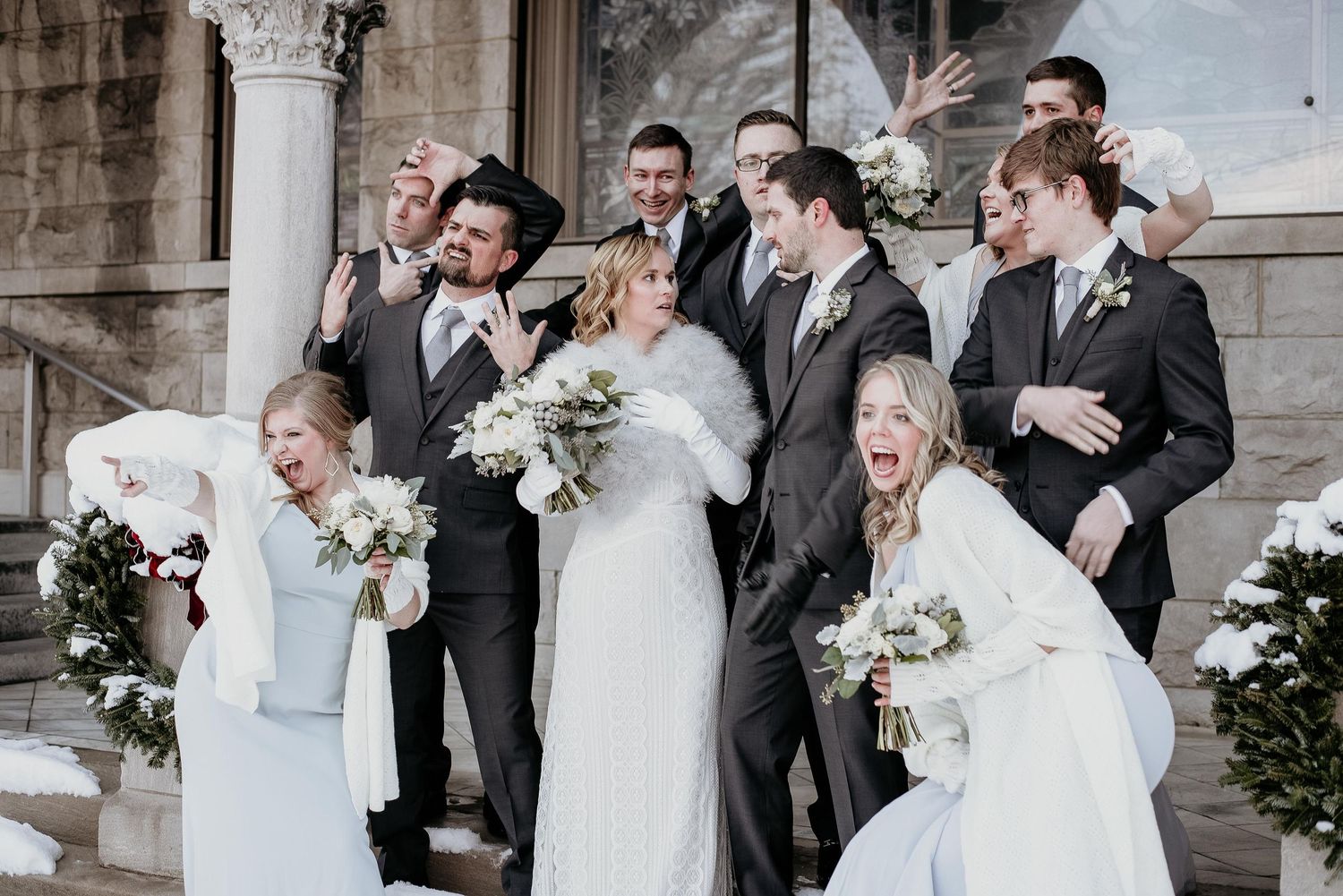 Wedding party poses playfully on snowy steps, bridesmaids in white wraps and groomsmen in gray suits, bride holding bouquet.