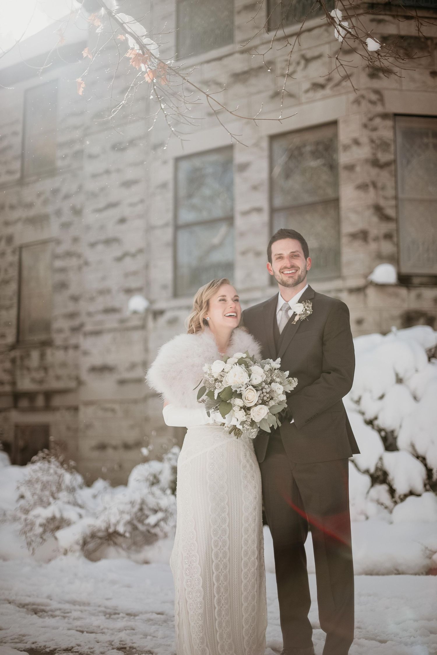 Smiling bride and groom pose in snow outside stone building, bride holding white bouquet wearing fur wrap over wedding dress.