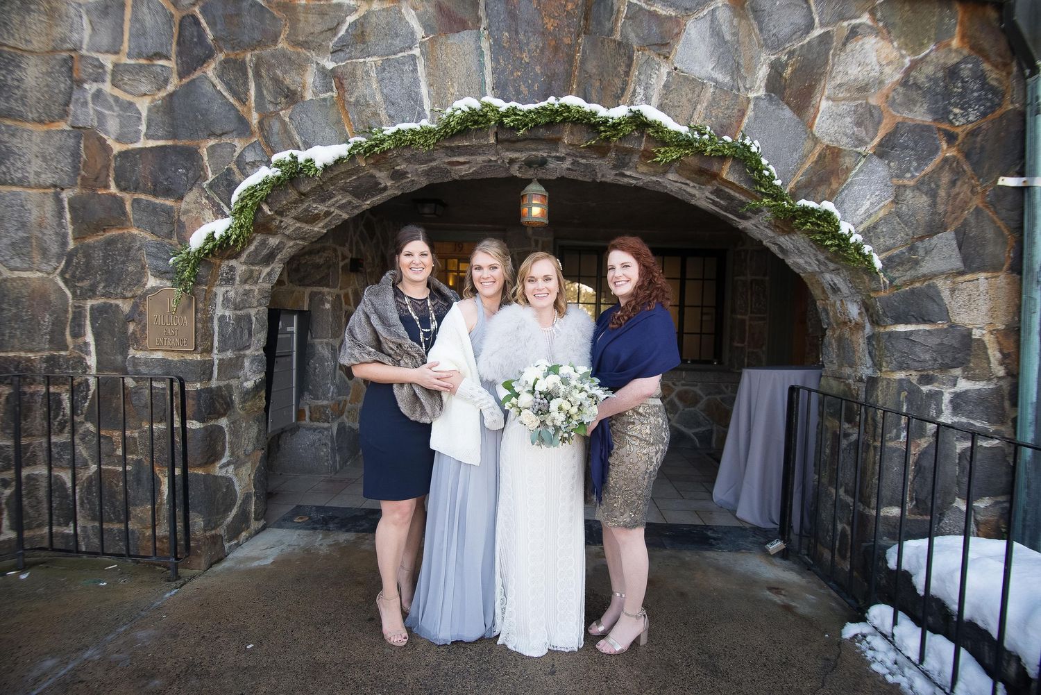 Bride in white gown poses with three bridesmaids under stone arch decorated with greenery at winter wedding venue.