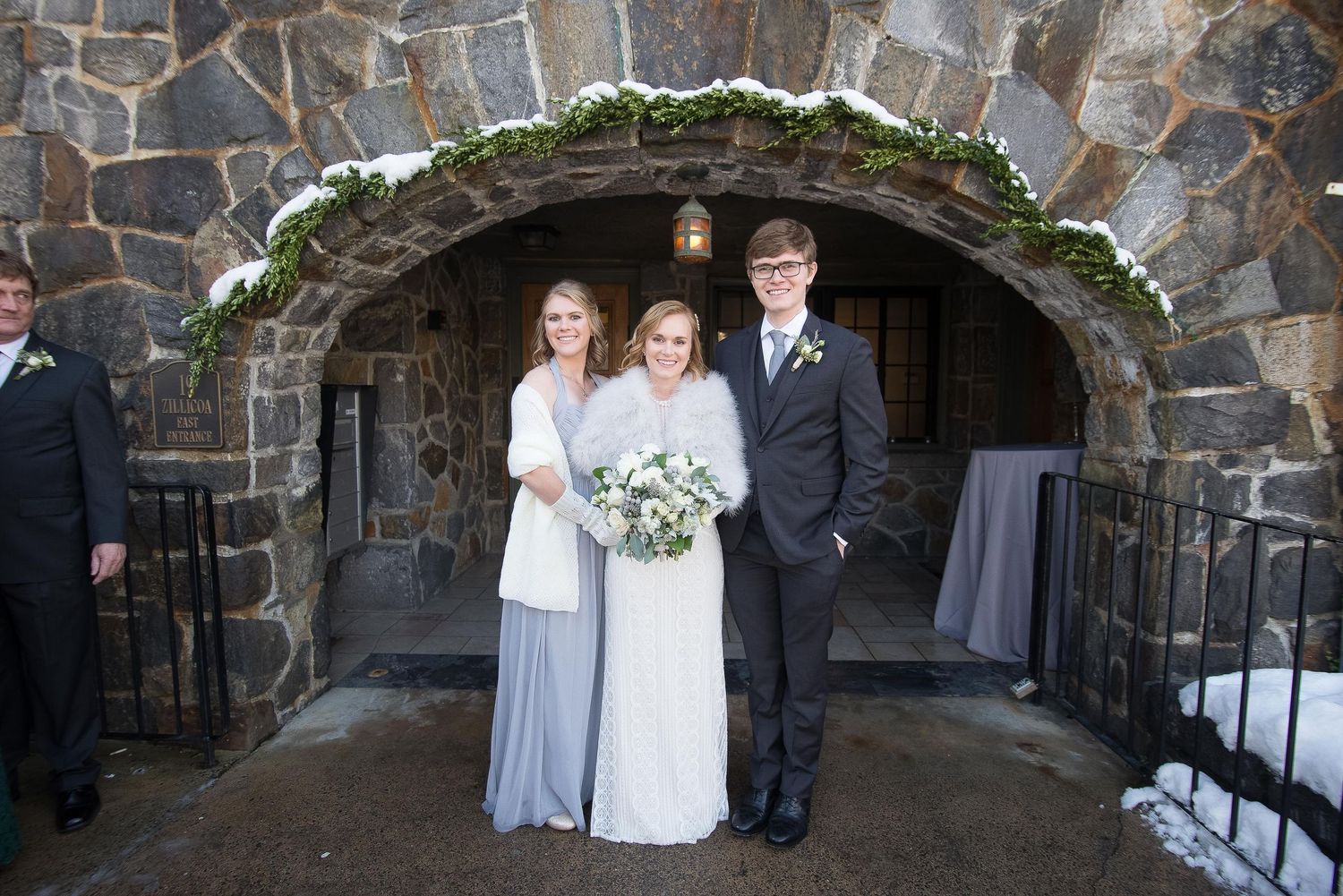 Bride in white gown poses with bridesmaid and groomsman under snowy stone arch at winter wedding venue.