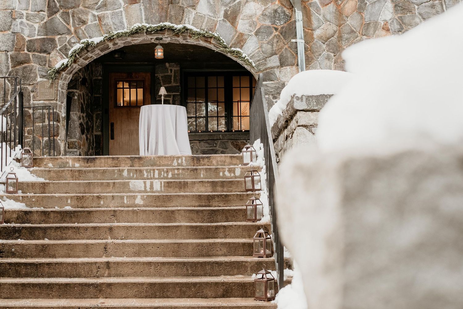 Snow-covered stone staircase leading to an elegant arched entrance with a draped table, perfect for a winter wedding venue.