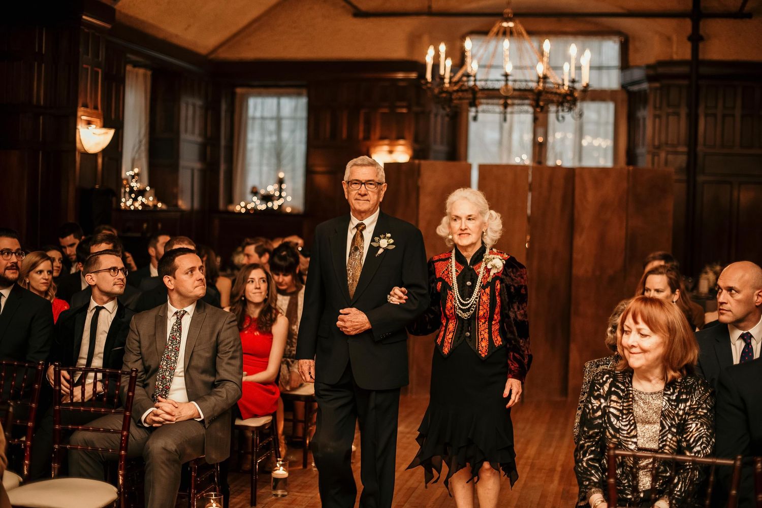 Elderly couple walking down the aisle at a wedding ceremony in an elegant wood-paneled room with seated guests.