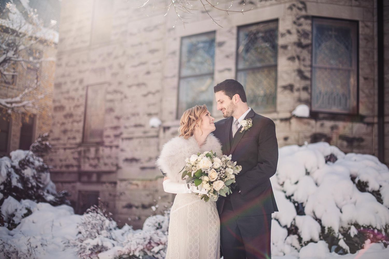 Bride and groom embrace in snowy winter wedding portrait outside stone building with bouquet of white roses.