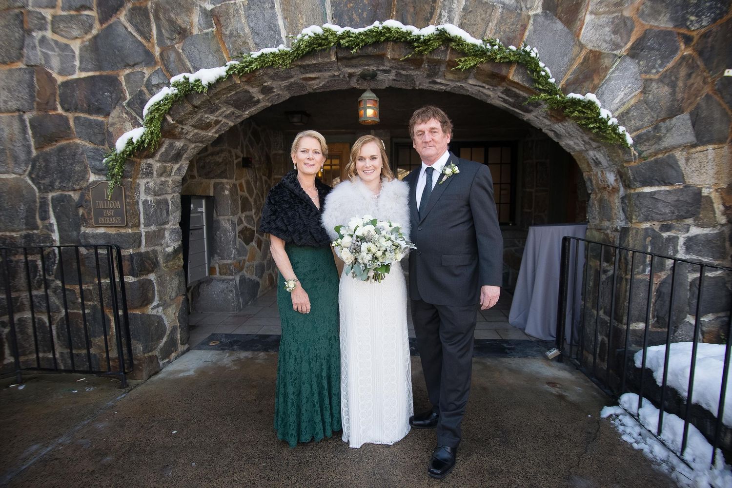 Bride in white gown poses with family in front of stone archway decorated with winter greenery.