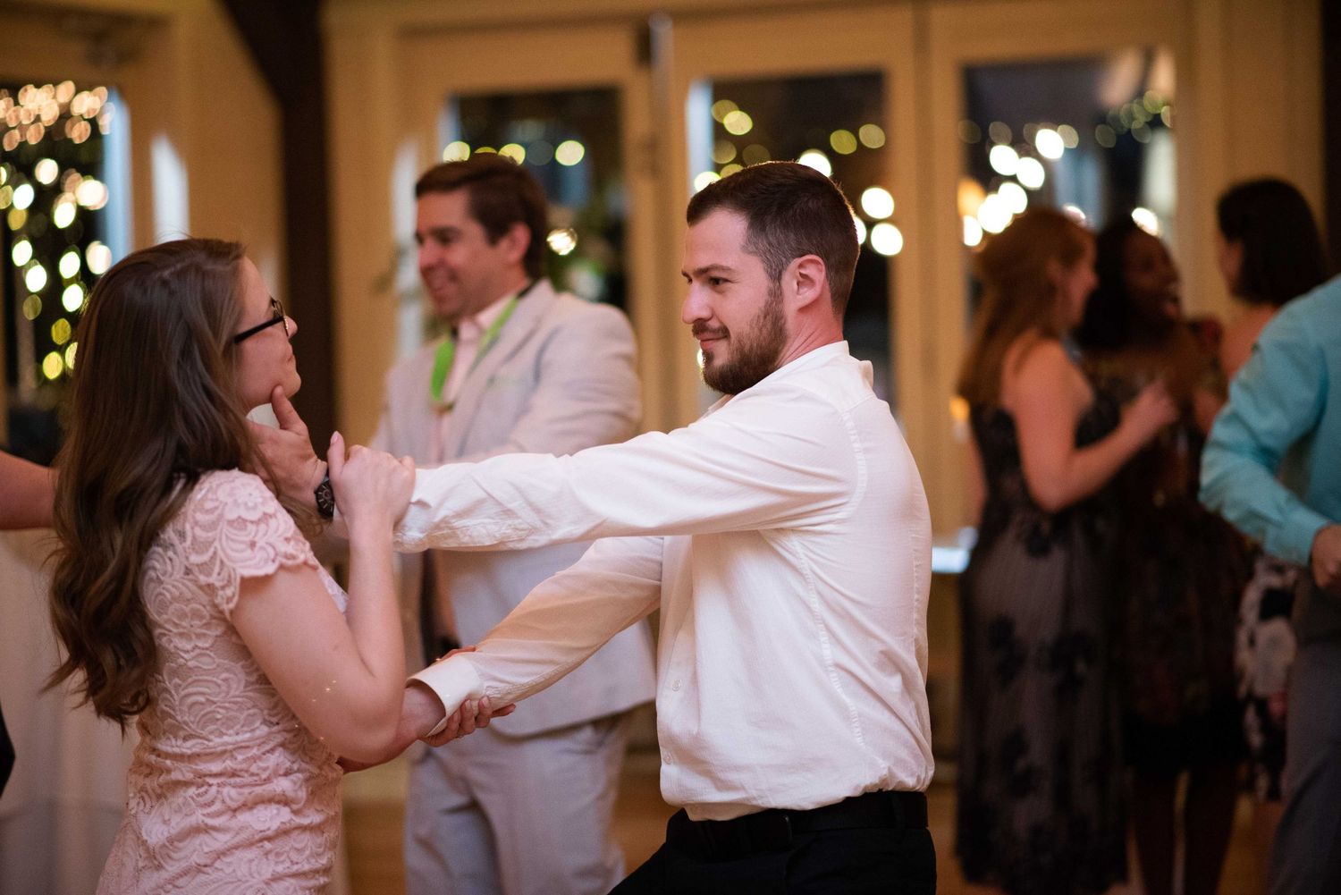 Couple dancing together at a wedding reception with guests in the background and warm string lights.
