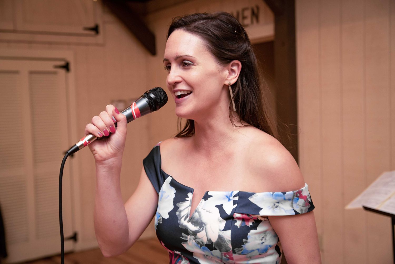 Woman in floral off-shoulder dress singing into microphone at indoor event.