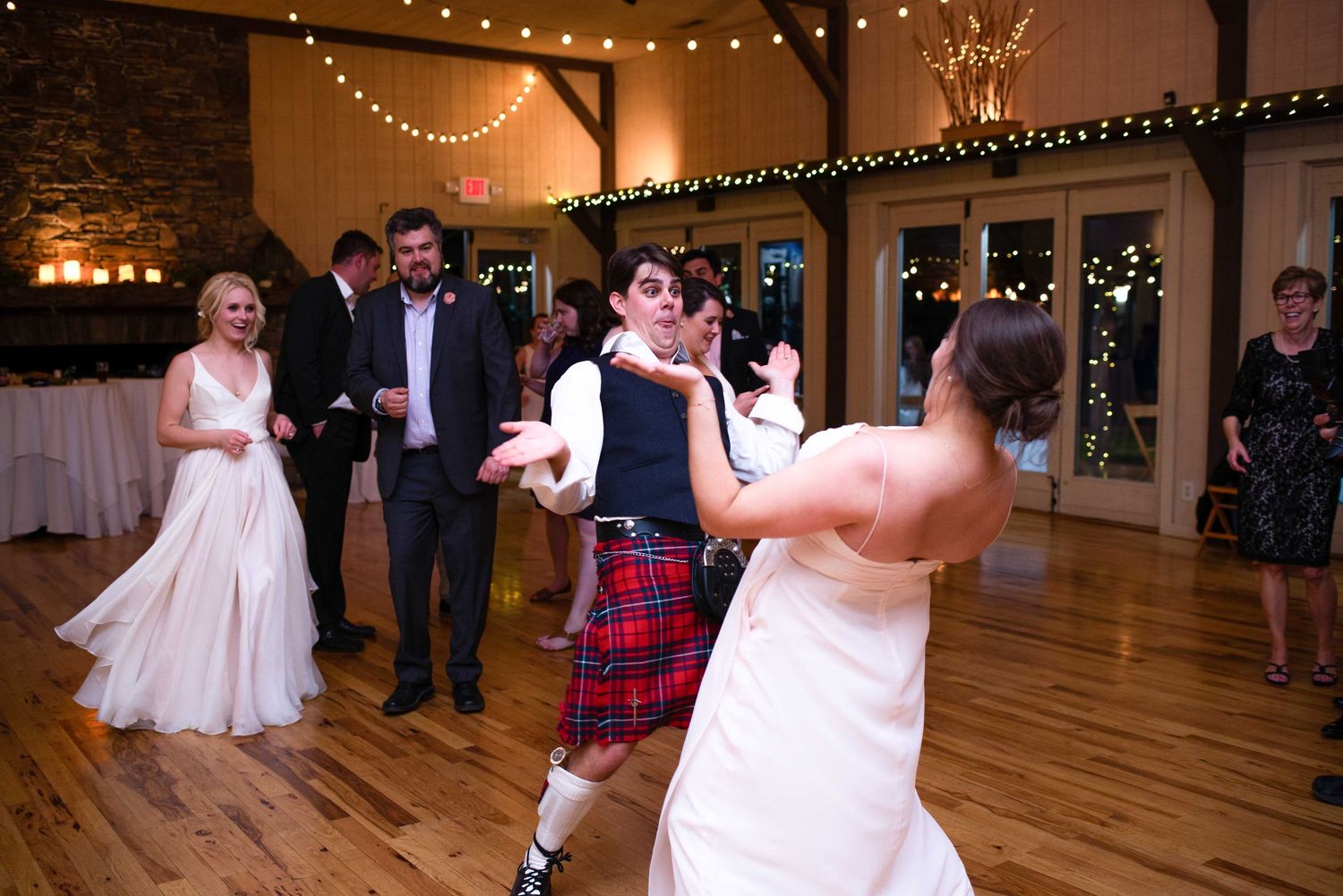 Bride and groom in Scottish kilt dancing joyfully with guests at elegant rustic wedding reception venue.