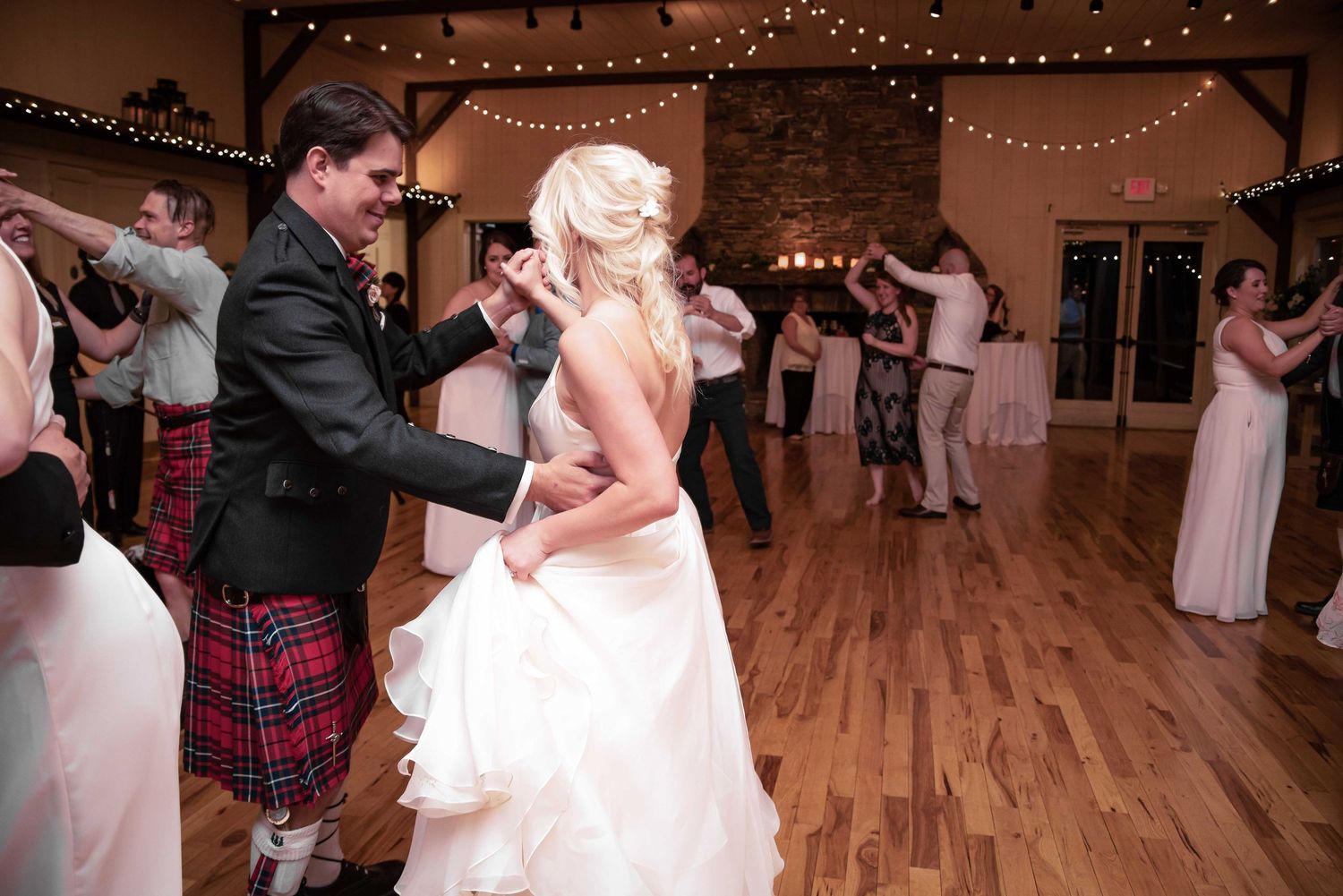 Bride and groom dancing together at wedding reception on wooden dance floor with guests and string lights.