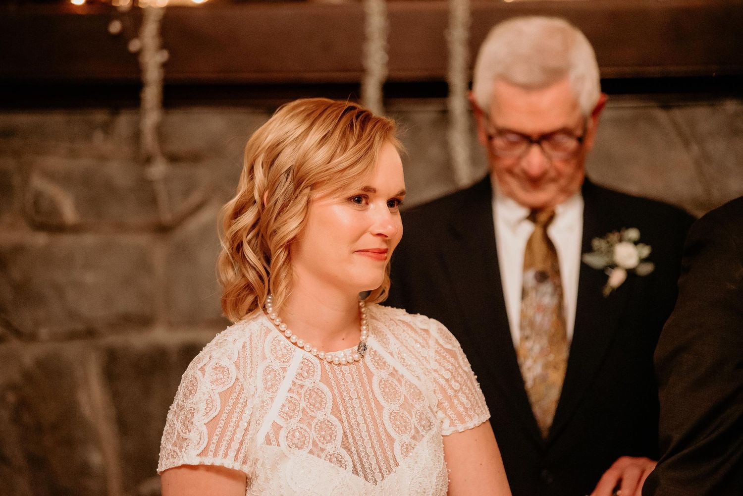 Bride in white lace dress smiling during wedding ceremony with officiant in background.