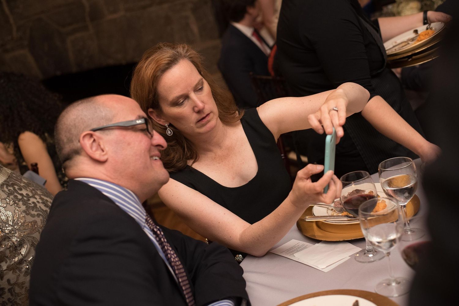 Man and woman at formal dinner table, woman holding phone showing something to seated man in glasses and tie.