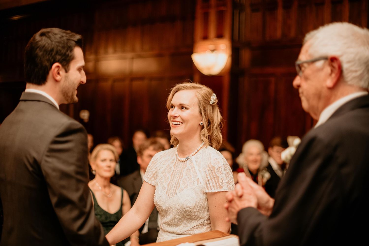 Bride and groom smiling during indoor wedding ceremony with guests seated in elegant wood-paneled venue.