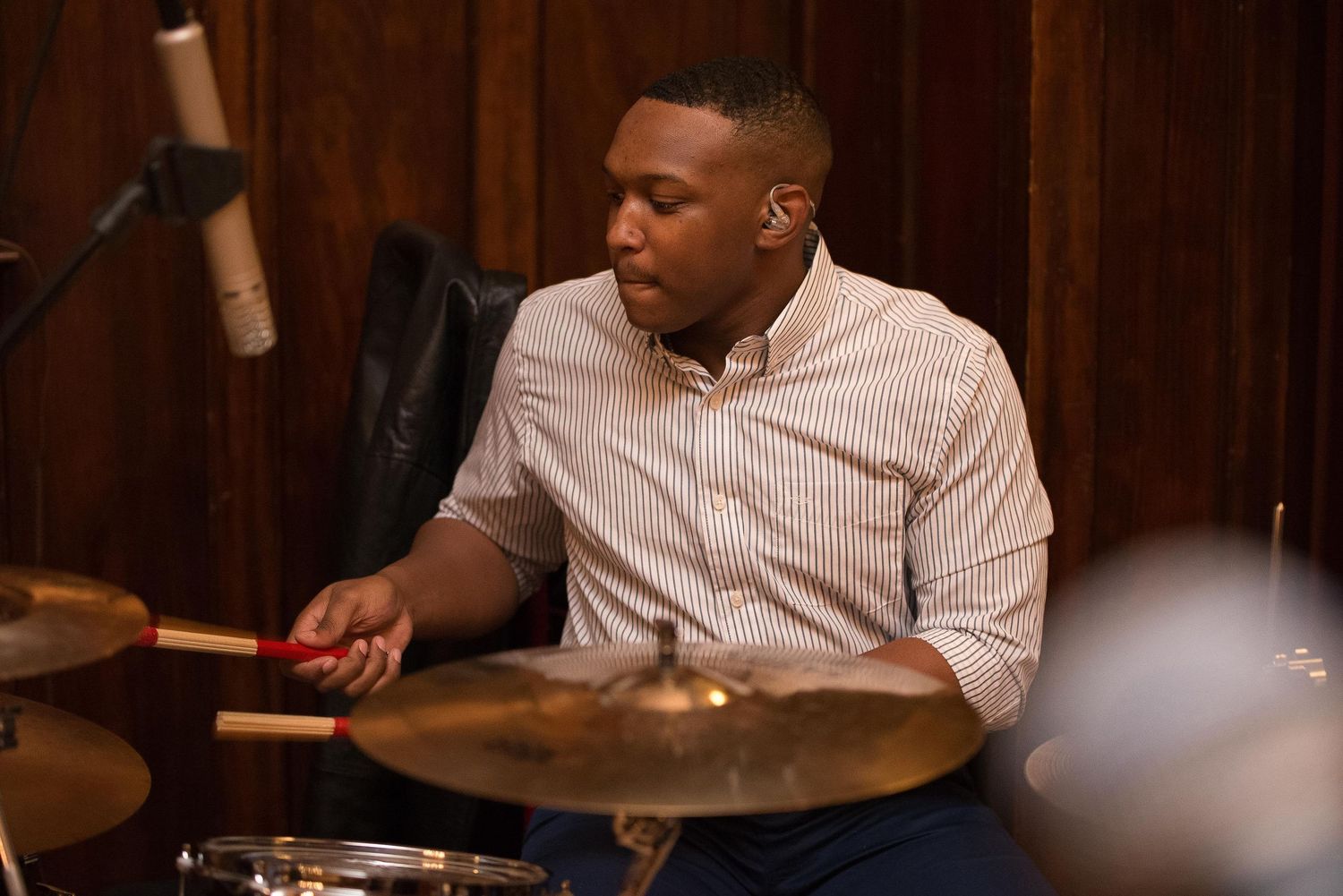 A young man plays drums with sticks in a recording studio, wearing a striped shirt, surrounded by cymbals.