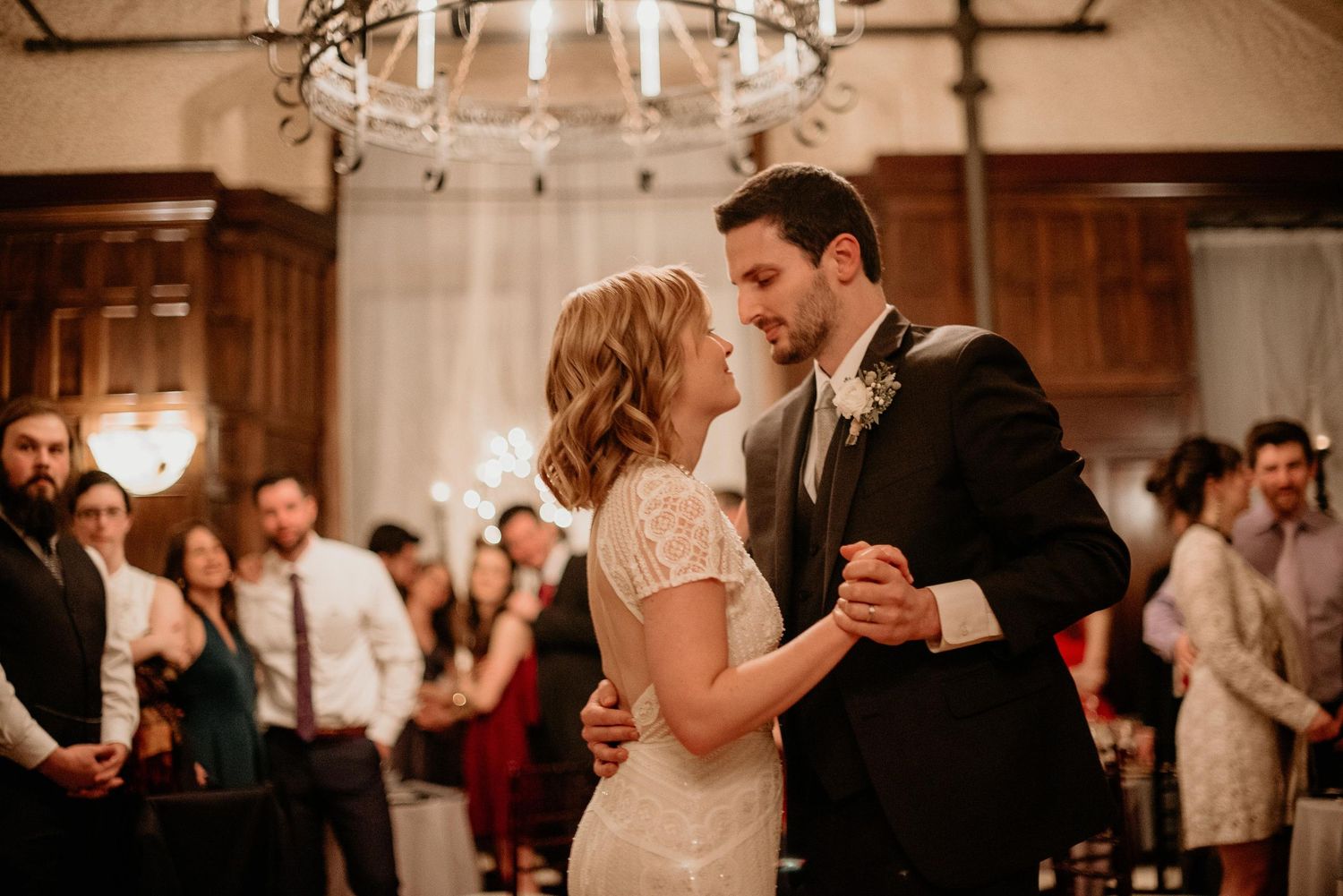 Bride and groom share first dance at elegant wedding reception with chandelier and guests watching.