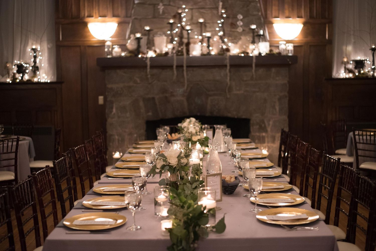 Elegant wedding reception table with gold plates, candles, greenery centerpieces, and a stone fireplace backdrop.