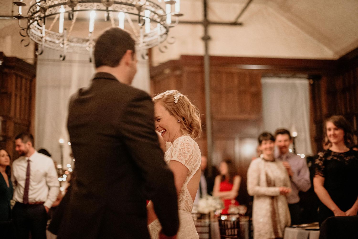 Bride laughing joyfully during first dance with groom in elegant wood-paneled reception hall with chandelier.