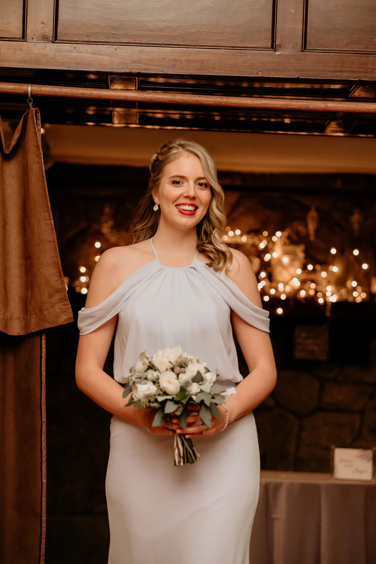 Smiling bride in off-shoulder gray gown holds white floral bouquet before a warmly lit fireplace backdrop.