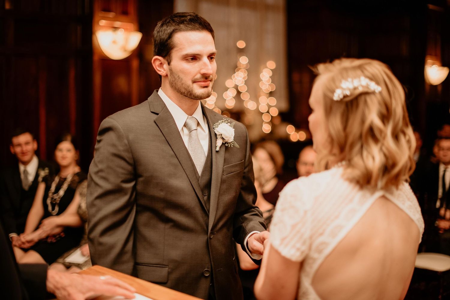 Groom in gray suit exchanges vows with bride during indoor wedding ceremony with warm bokeh lights.