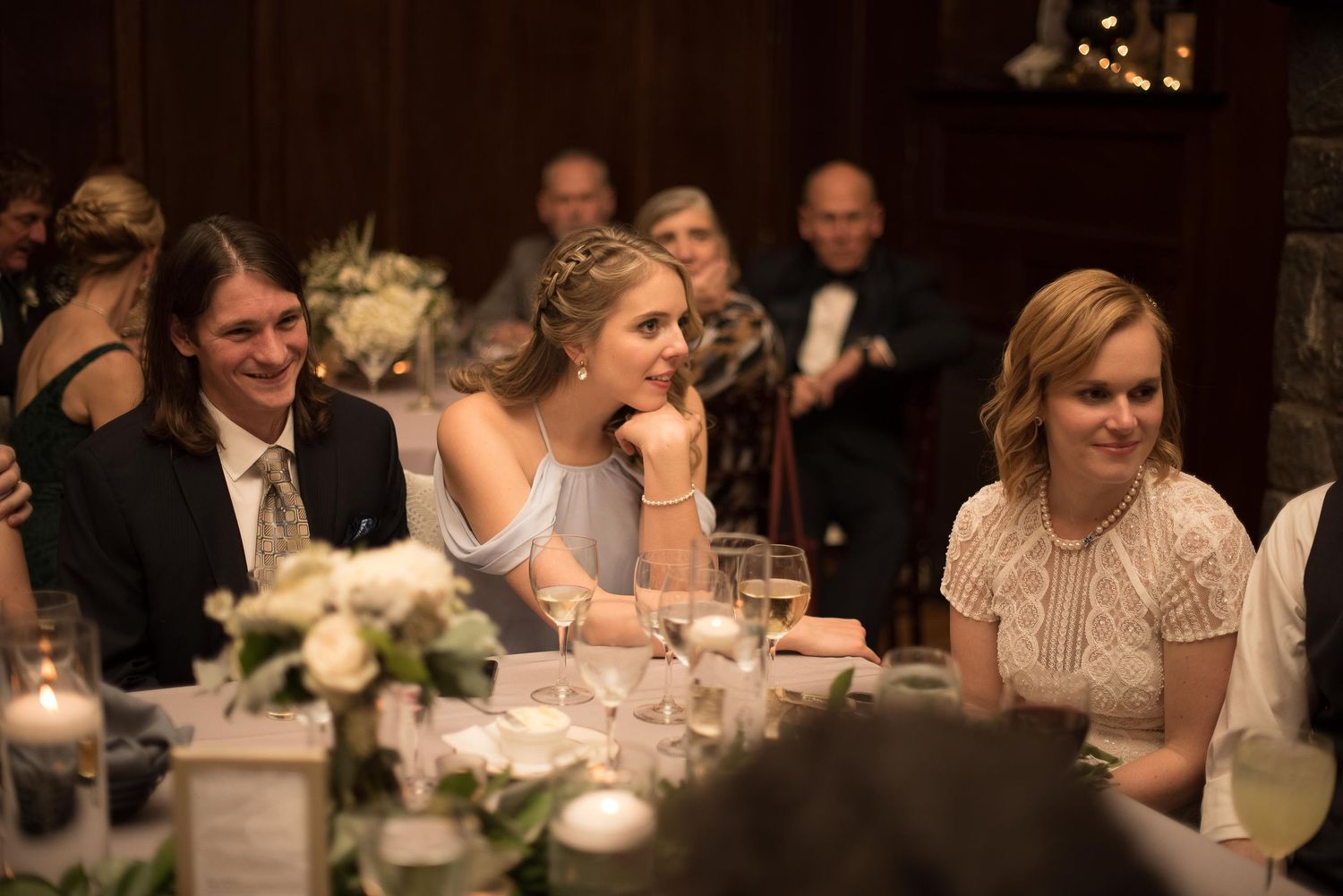 Wedding guests seated at reception table smiling during speeches at formal evening event.