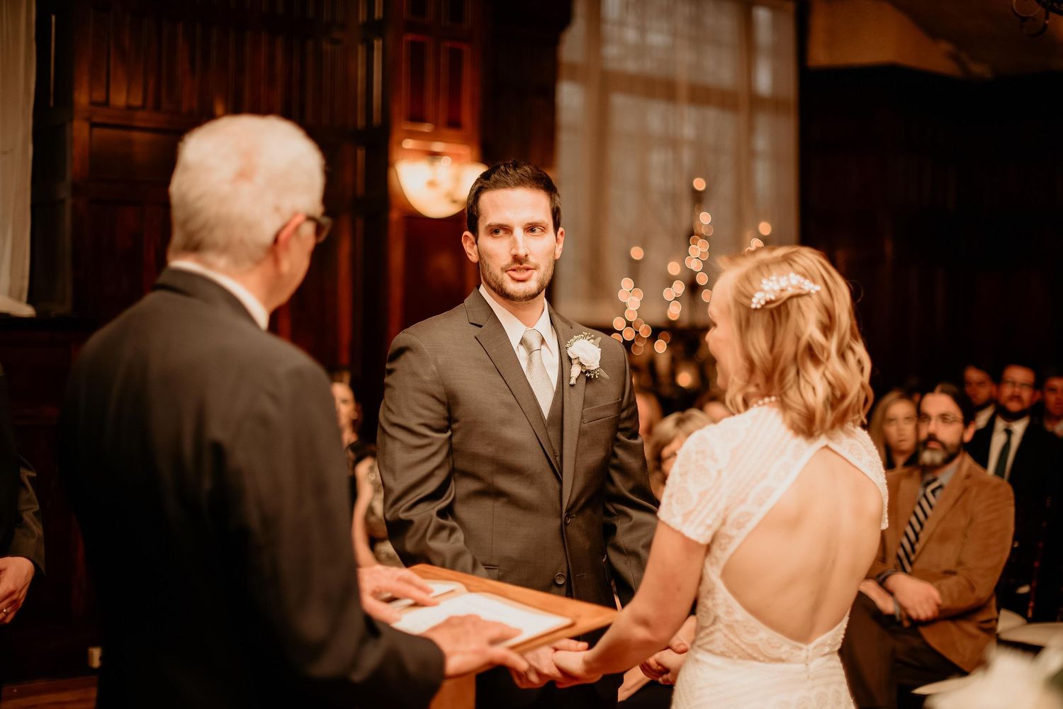 Groom smiling at bride during indoor wedding ceremony, officiant holding book, warm romantic lighting.