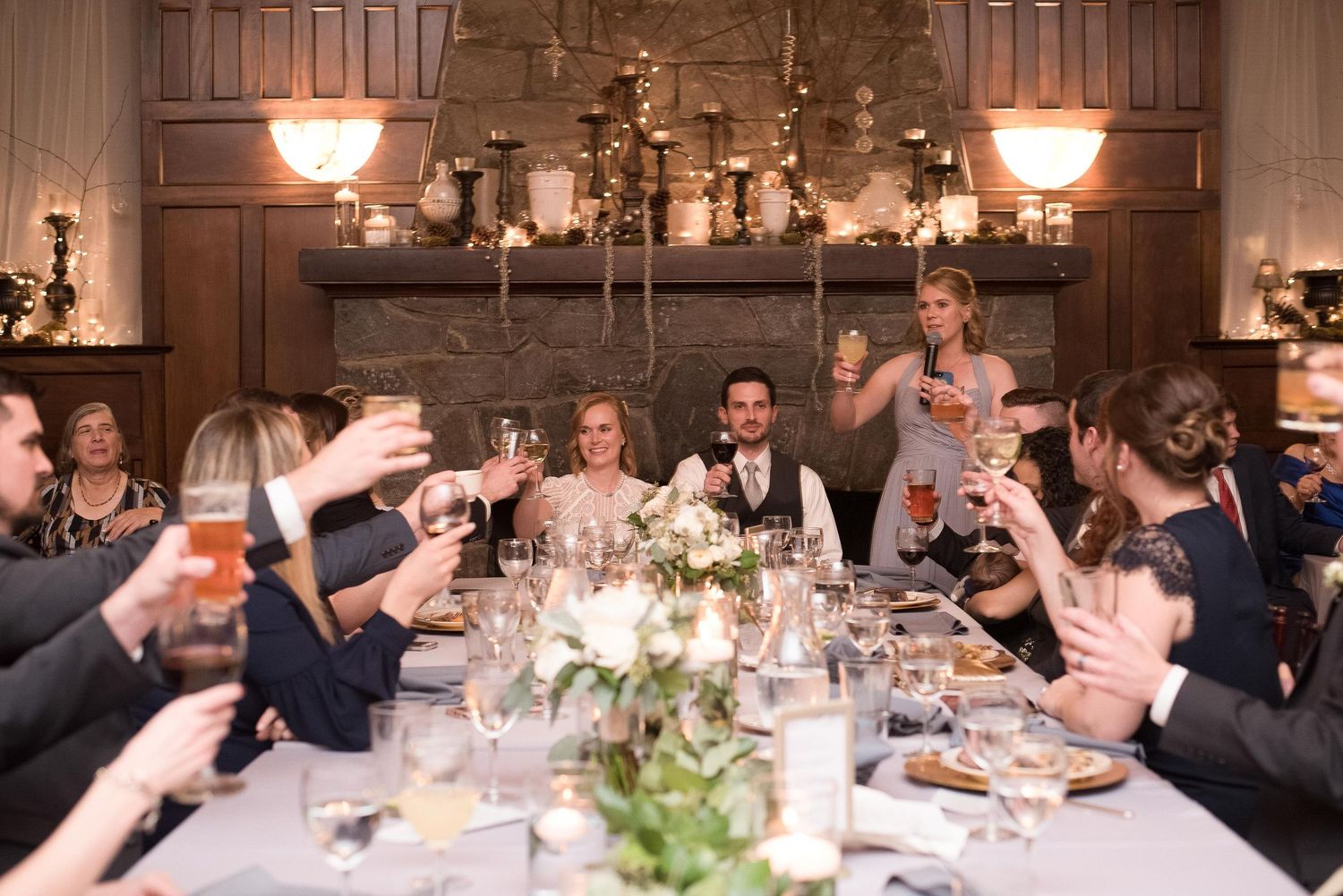 Elegant wedding reception toast with guests raising glasses around a candlelit table in a warmly lit dining room.