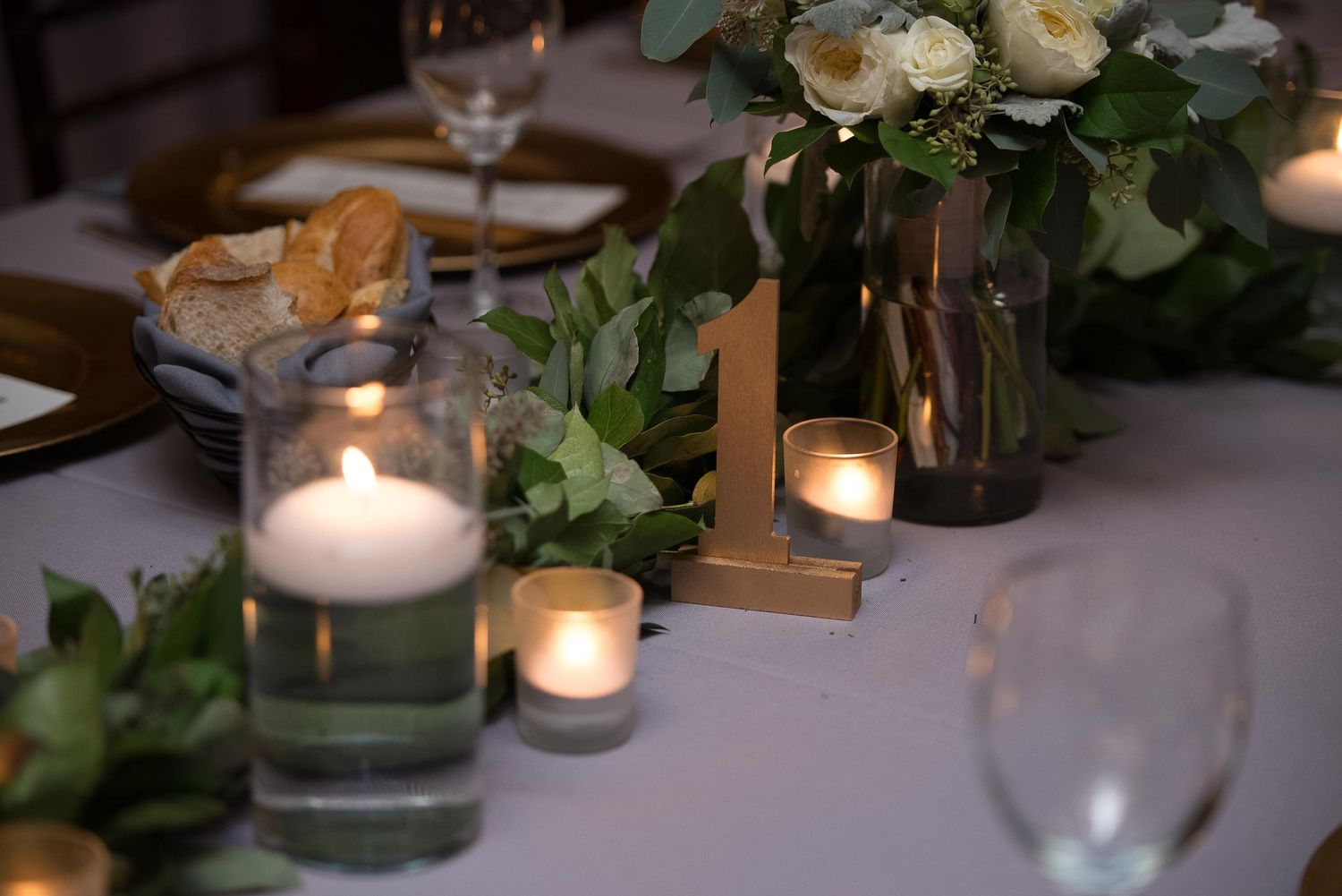 Elegant wedding table setting with white candles, greenery, gold table number, and white floral centerpiece.