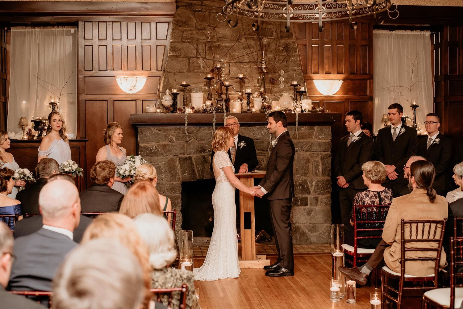 Couple exchanges vows indoors at rustic wedding ceremony with stone fireplace, candlelight, and seated guests.