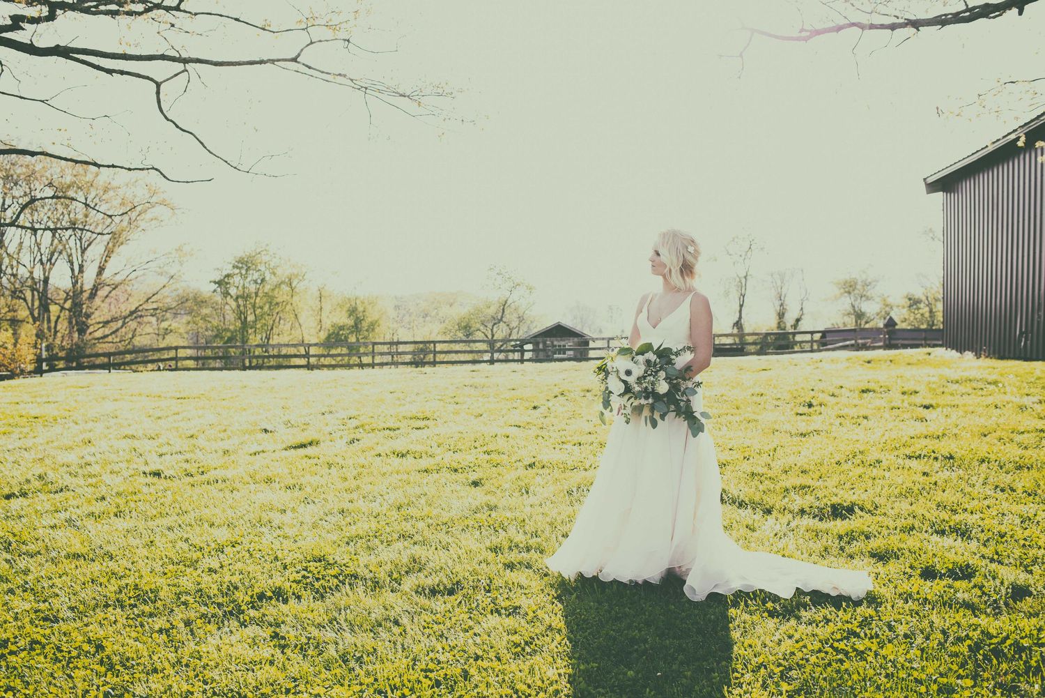 Bride in white gown holding bouquet stands in lush green field near rustic barn on sunny wedding day.