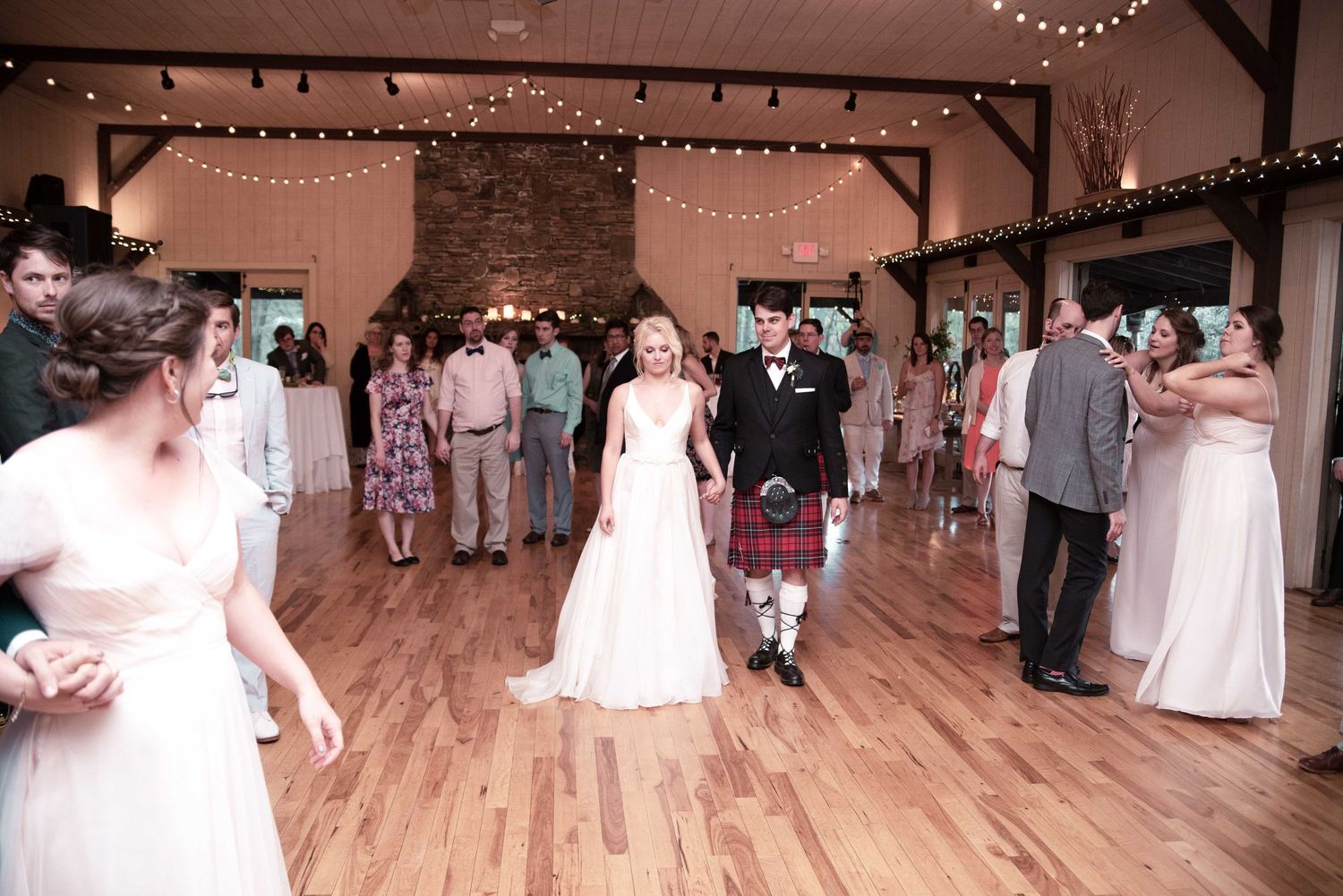 Bride and groom in Scottish kilt walk hand-in-hand at rustic barn wedding reception with string lights.