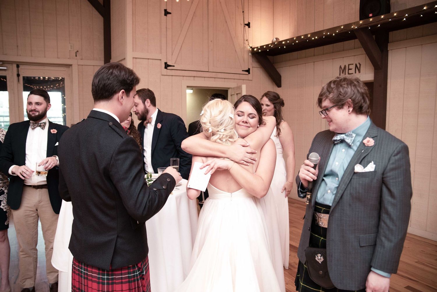 Bride in white gown hugging guest at rustic barn wedding reception with groomsmen in kilts nearby.