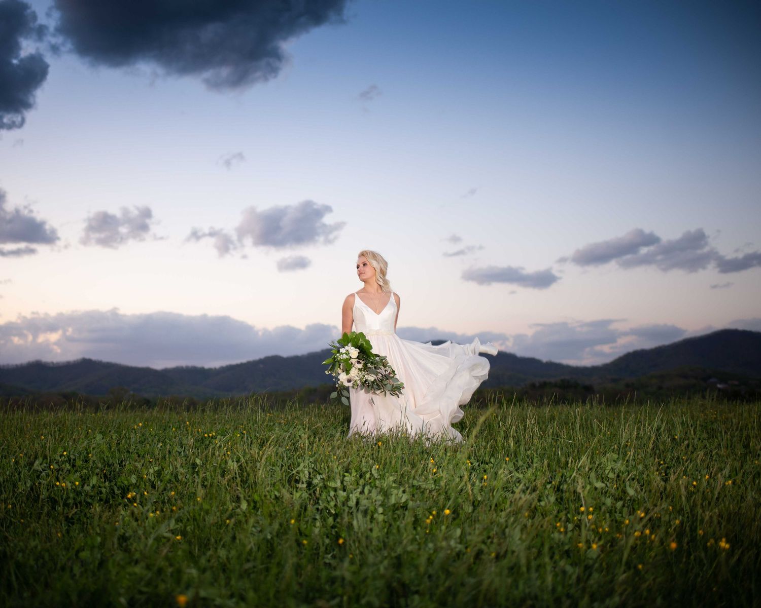 Bride in flowing white gown holding bouquet stands in green meadow with mountain backdrop at sunset.