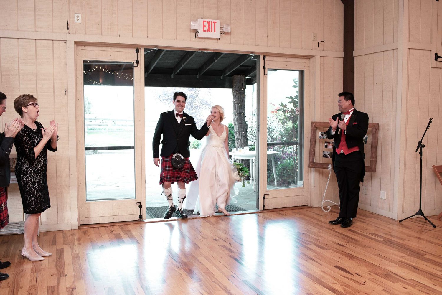 Bride and groom make grand entrance at wedding reception, groom wearing traditional Scottish kilt, guests applauding.