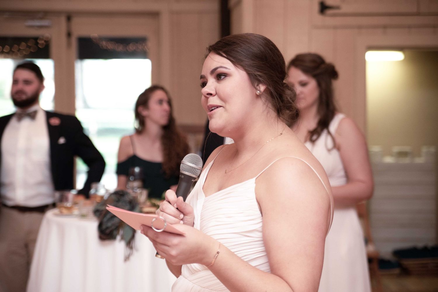 Woman in white dress giving a speech at a wedding reception while holding a microphone and note cards.