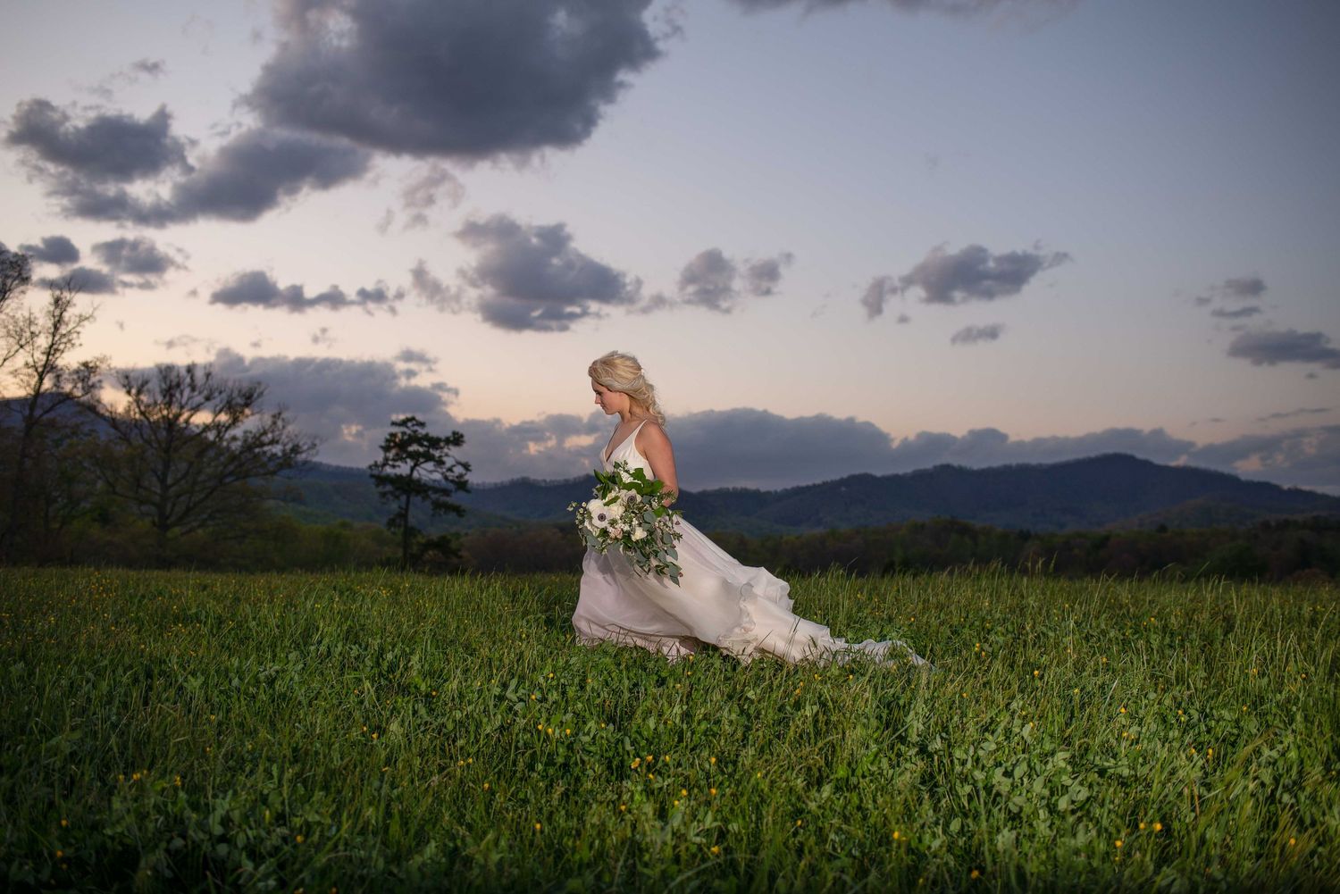 Bride in flowing gown walks through green field at dusk with mountain backdrop and bouquet.