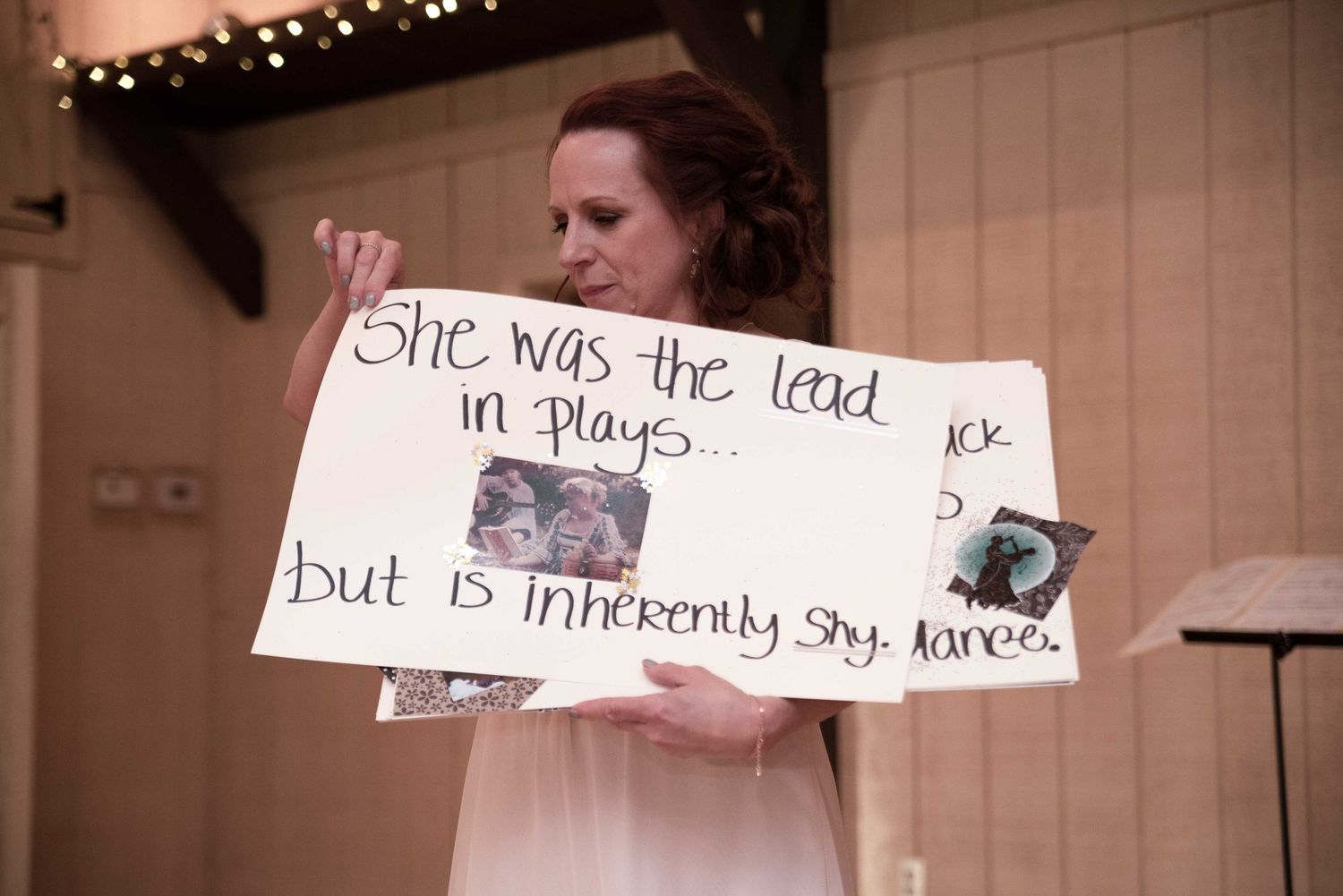 Woman holding handwritten sign reading 'She was the lead in plays but is inherently shy' at a wedding reception.
