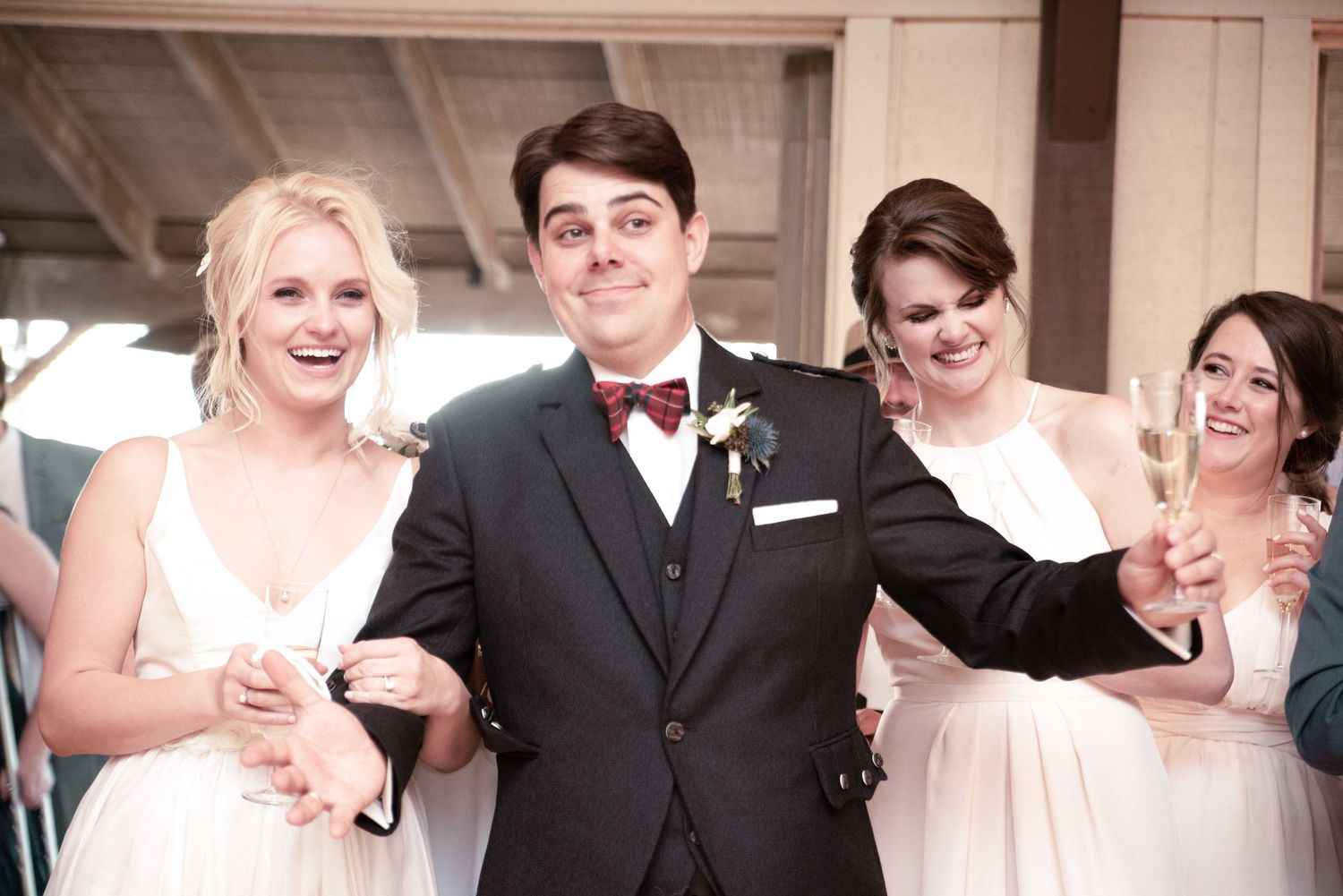 Groom in black tuxedo with bow tie smiles with bridesmaids in white dresses at wedding reception celebration.